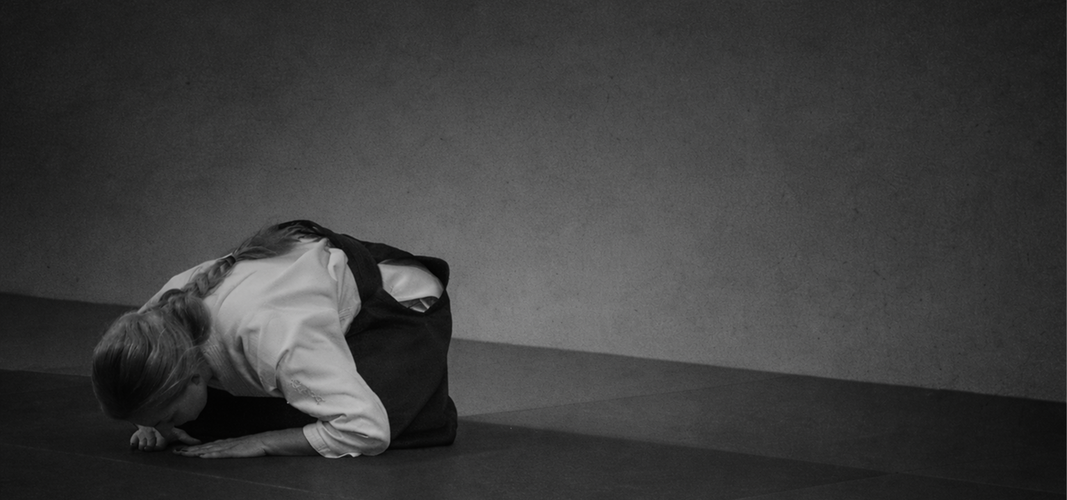 A woman in a martial arts uniform in a kneeling position and bowing on the floor in an indoor training area.
