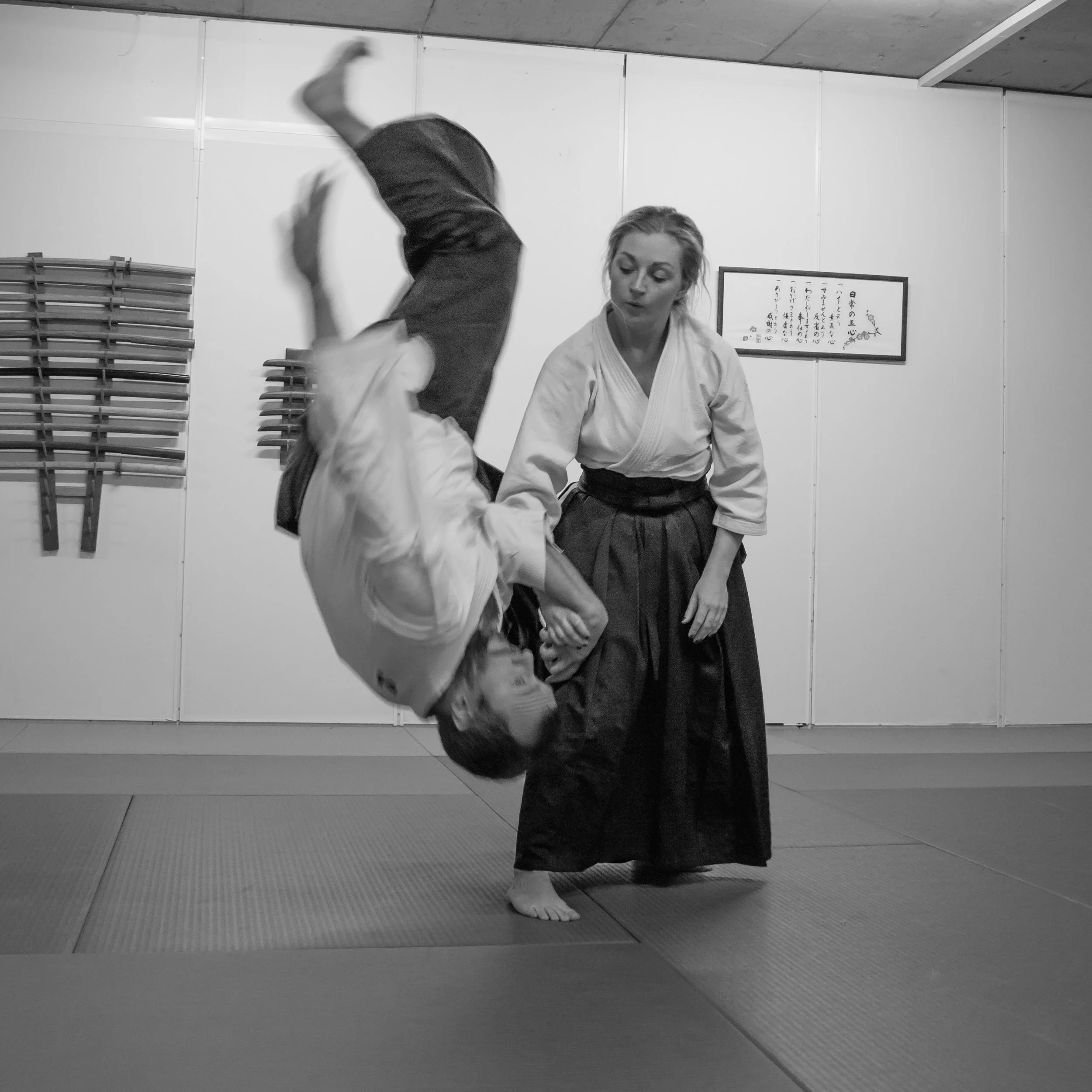 Two women practicing Aikido in a dojo, one performing a throw on the other while the other observes.