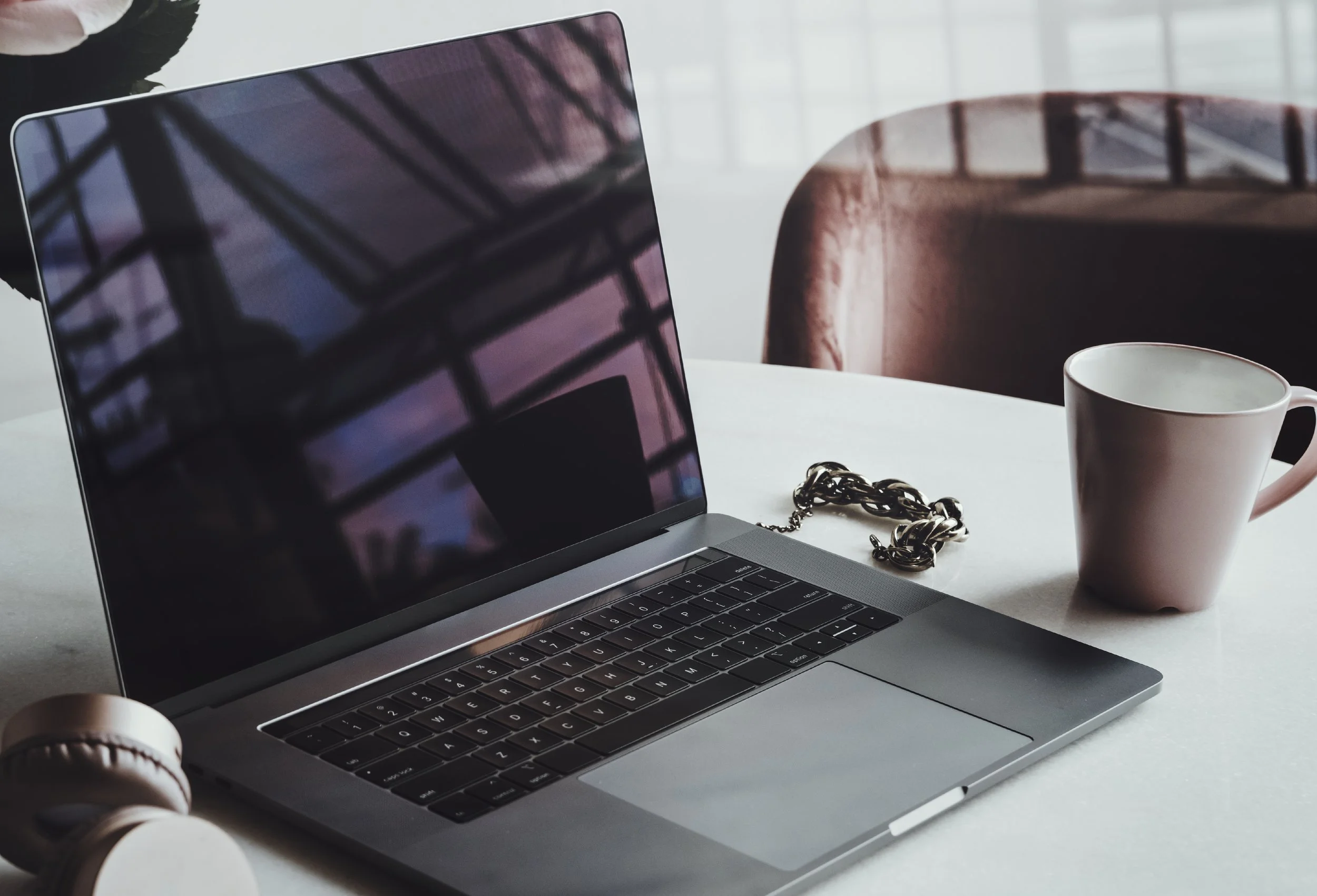 A laptop, a white coffee mug, a chunky silver chain, and a pair of beige headphones on a table, with a brown chair and a window in the background.