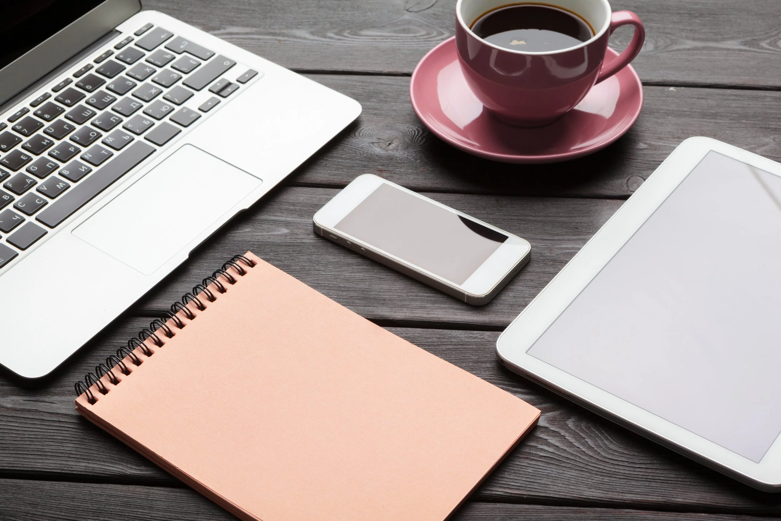 A laptop, a smartphone, a tablet, a pink notebook, a pink cup of coffee on a saucer on a wooden table.