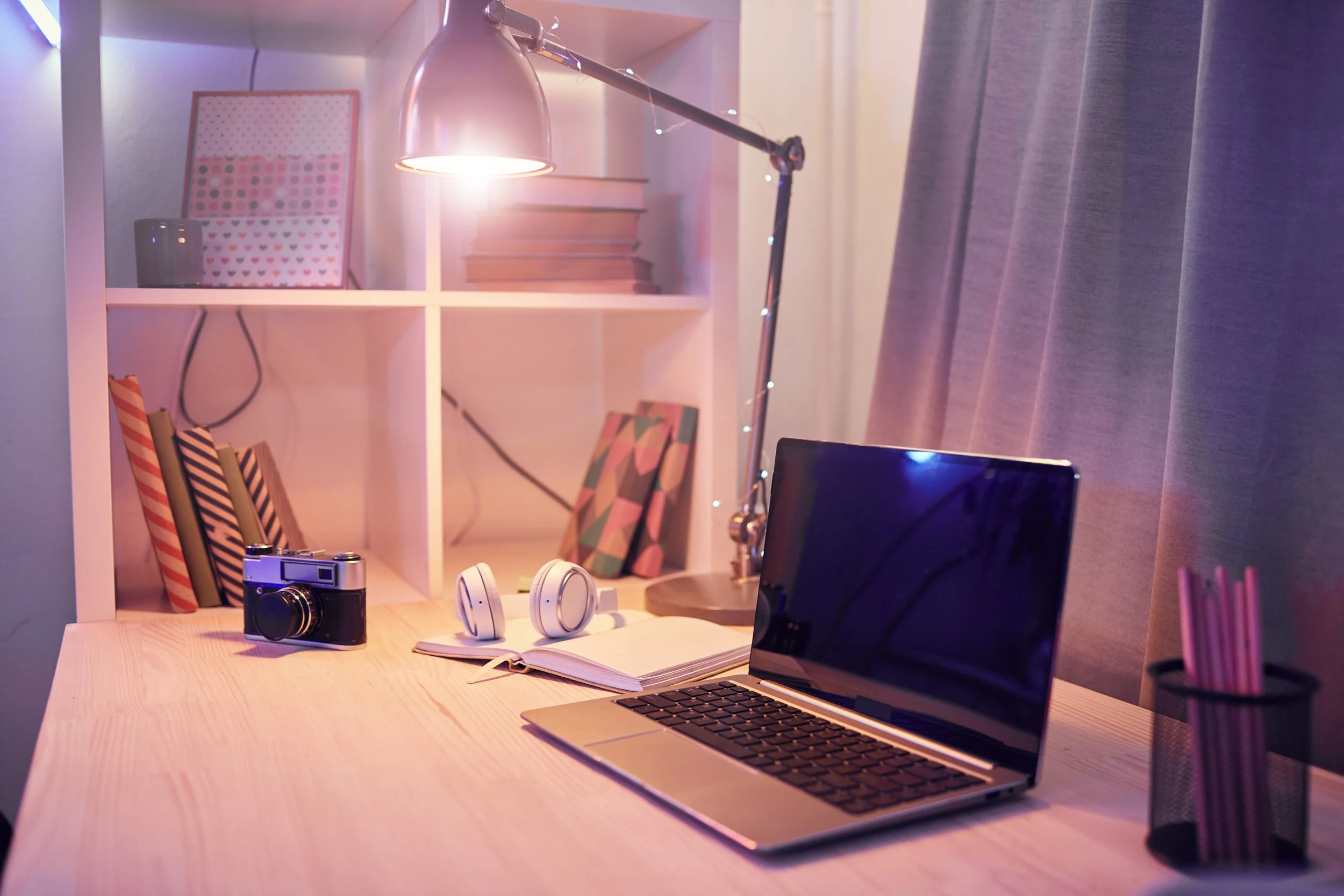 A tidy desk with a laptop, headphones, a camera, an open notebook, pink pencils in a black holder, and a desk lamp. Behind the desk is a white shelf with books, a framed planner, and decorative object, with pink and colorful notebooks under the shelf. The scene is lit with soft pink and purple lighting.