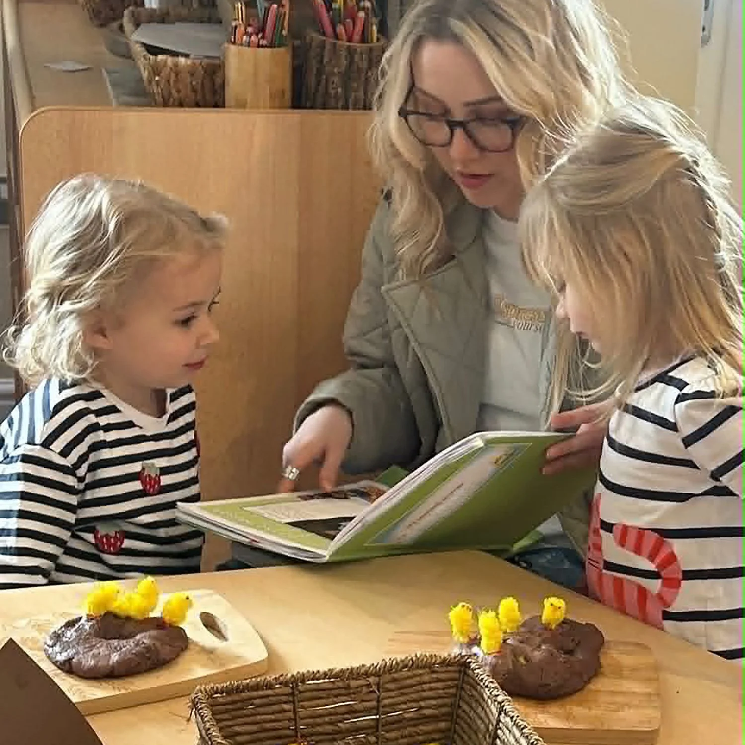 Two young girls and a woman looking at an open book together in a kitchen or dining area. A table in front has chocolate cookies decorated with yellow chicks, likely for Easter.