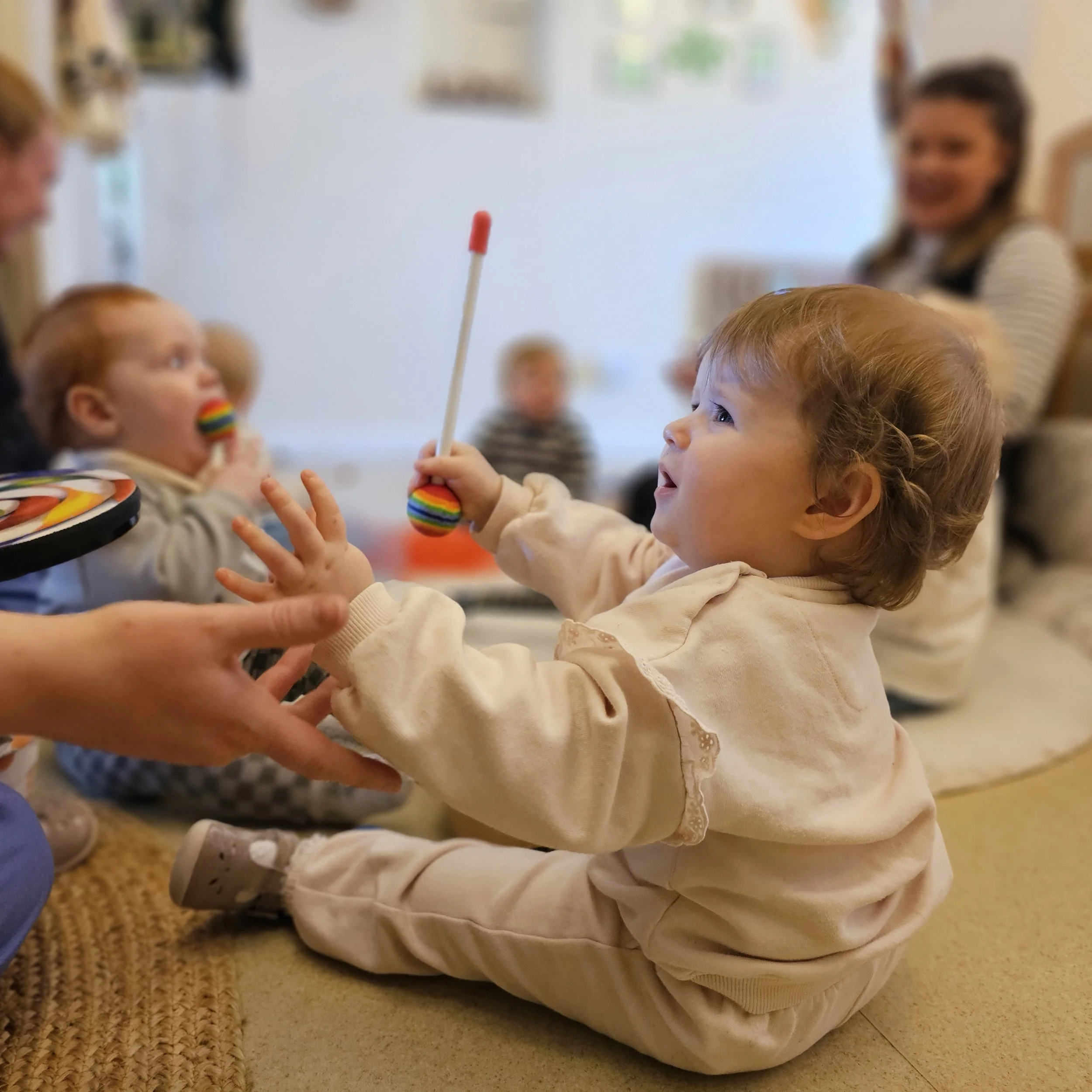 A close-up of a young child with curly hair, reaching out towards a person holding a bottle with a red tip, sitting on the floor next to a white bag and a colorful object, in an indoor setting with a carpeted floor and a blurred background with a woman and other children.