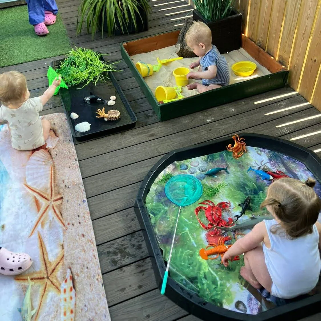 Children playing with toy ocean and beach-themed toys on a wooden deck, including a sandbox with sand toys, a water table with toy sea creatures, and a small tray with plants and shell decorations.