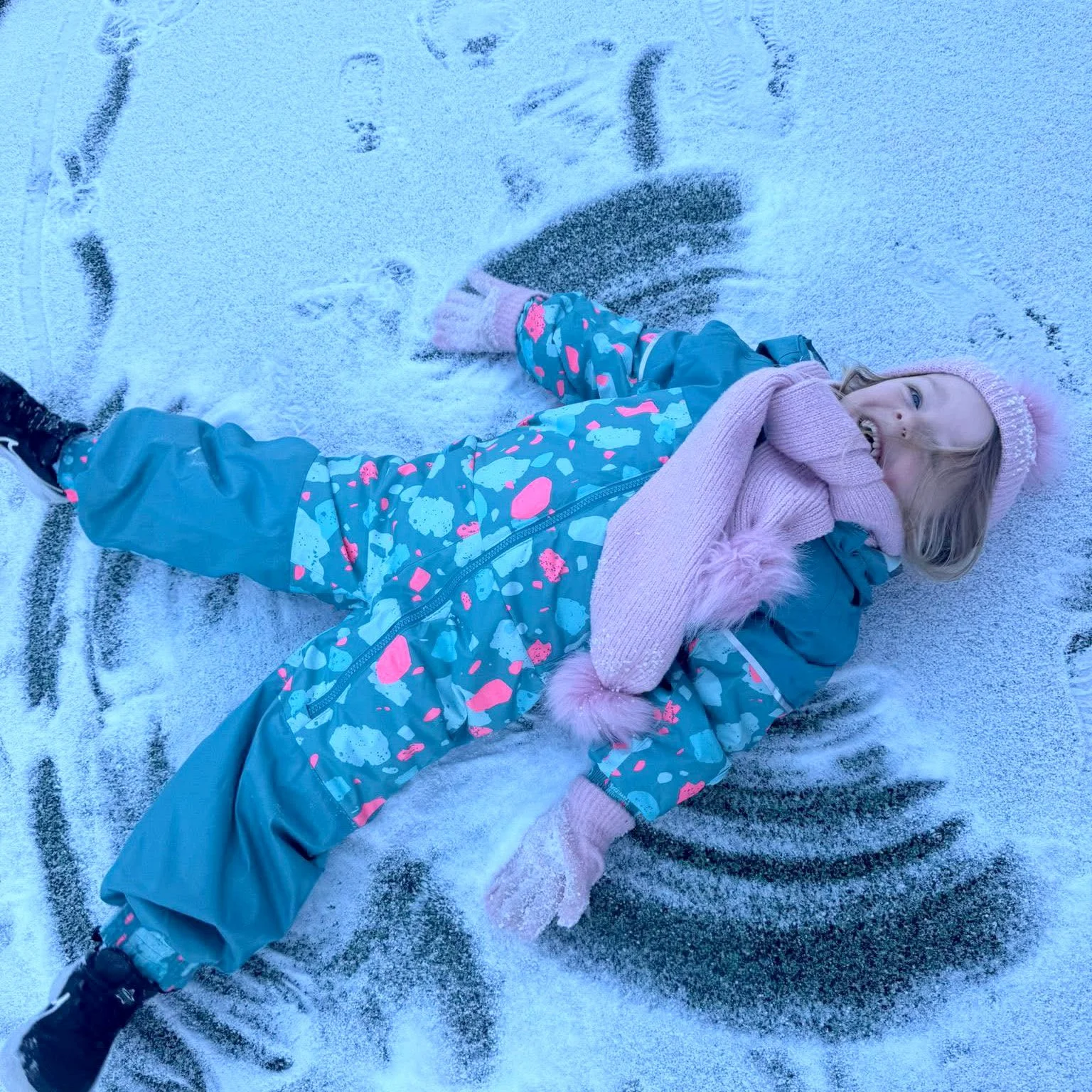 A young girl with blonde hair lying on snow, smiling, wearing a pink knit hat, pink gloves, a blue snowsuit with colorful patterns, and black boots.