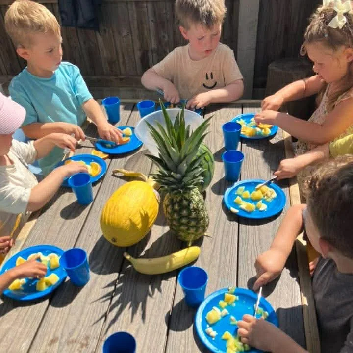 Children sitting around a wooden table enjoying a meal with yellow fruit pieces on blue plates, with pineapples, a melon, and bananas in the center, and blue cups.
