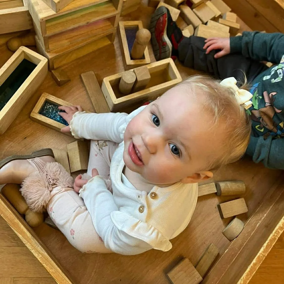 A young blonde child playing with wooden building blocks on a wooden floor, surrounded by scattered blocks, wearing a white long-sleeve shirt, pink leggings with floral patterns, and fuzzy slippers.