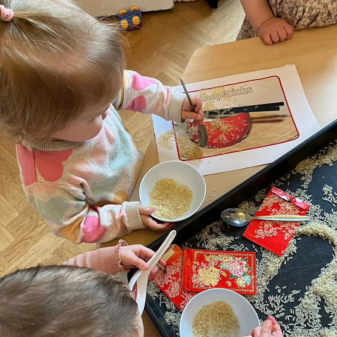 Children participating in a rice packaging activity, scooping rice into red packets and bowls, with scattered rice on a tray and a photo of a rice container in the background.