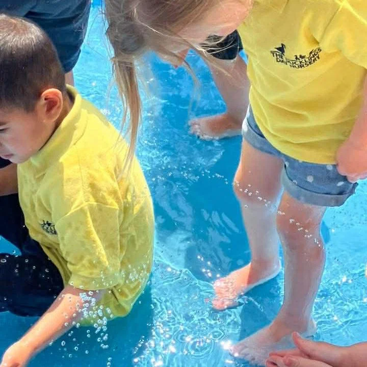 Two children and a person in a pool. The children are wearing yellow shirts, and the person is partially visible, likely an adult. All are standing in the shallow water.