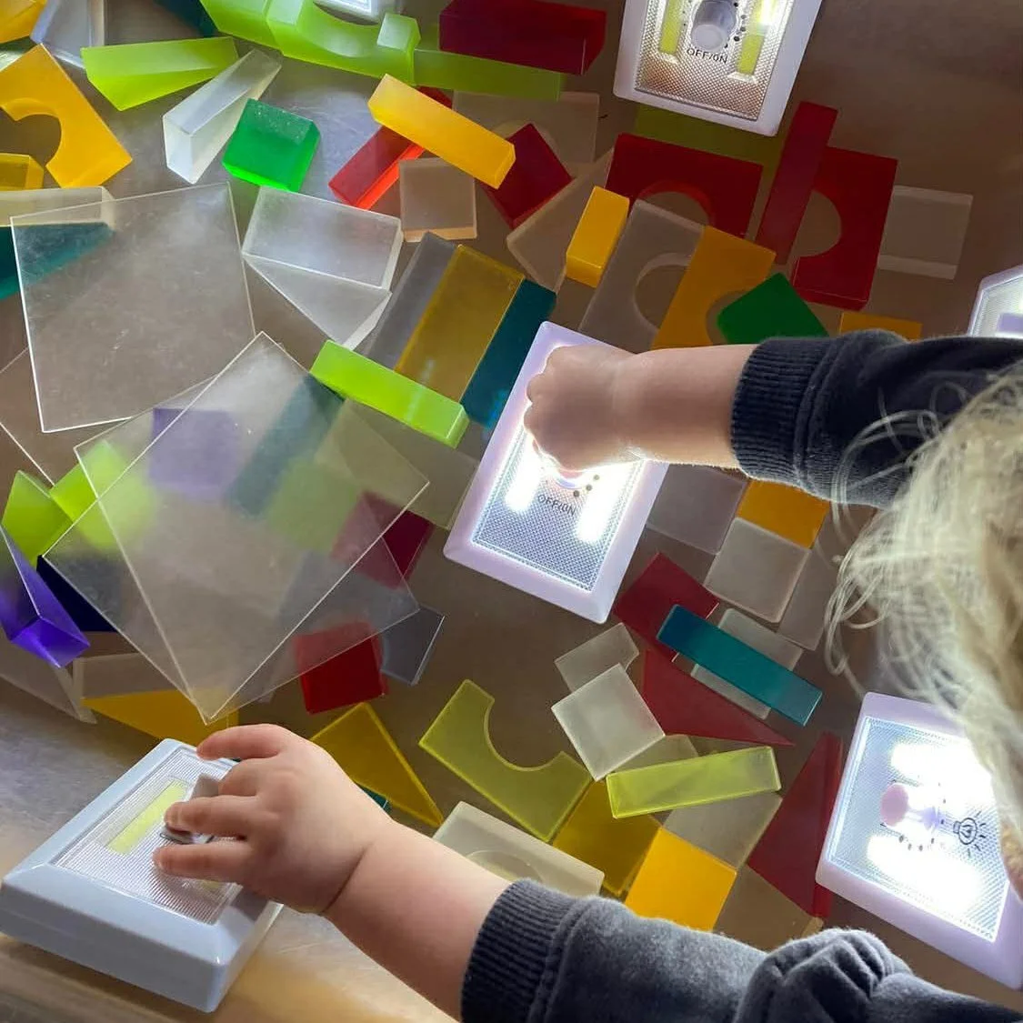 Children playing with colorful translucent geometric shapes and light boxes on a table.