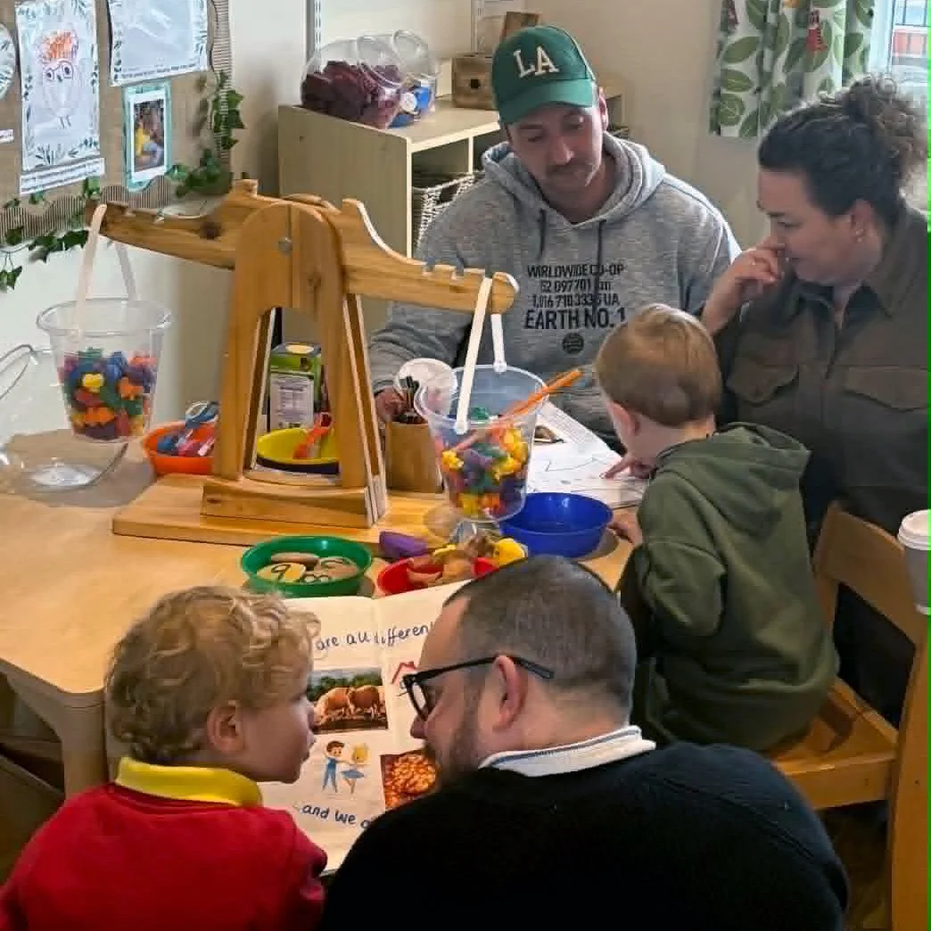 Adults and children gathered around a table engaged in a group activity, with colorful toys and artwork on display, in a cozy room with patterned curtains and children's pictures on the wall.