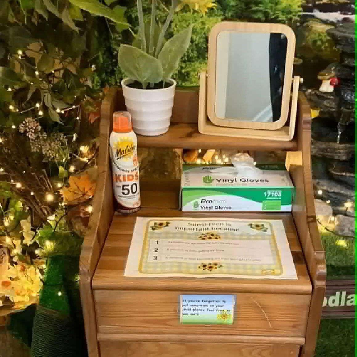 A small wooden table with a mirror, a potted plant, a bottle of sunscreen, a box of vinyl gloves, and a printed sign with a sunflower theme. The background includes decorative string lights and a stone wall.