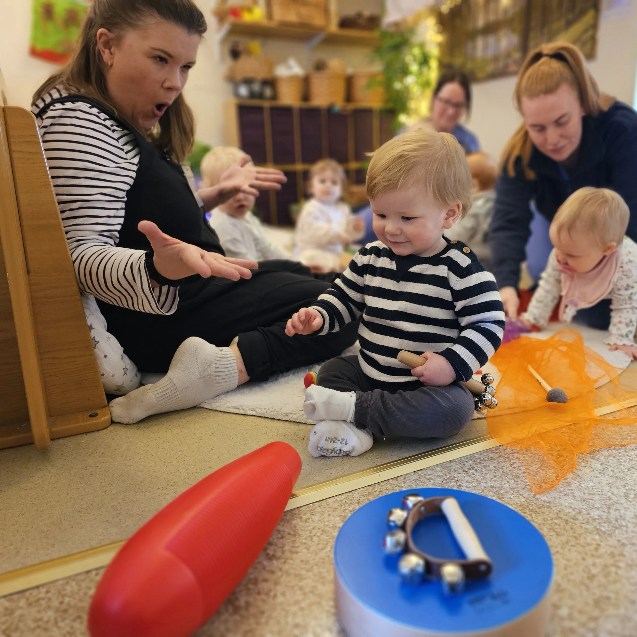 A classroom with children and teachers sitting on the floor. The focus is on a young boy with red hair in a striped shirt, sitting cross-legged, and a woman leaning over in the background. There are toys and colorful decorations around.