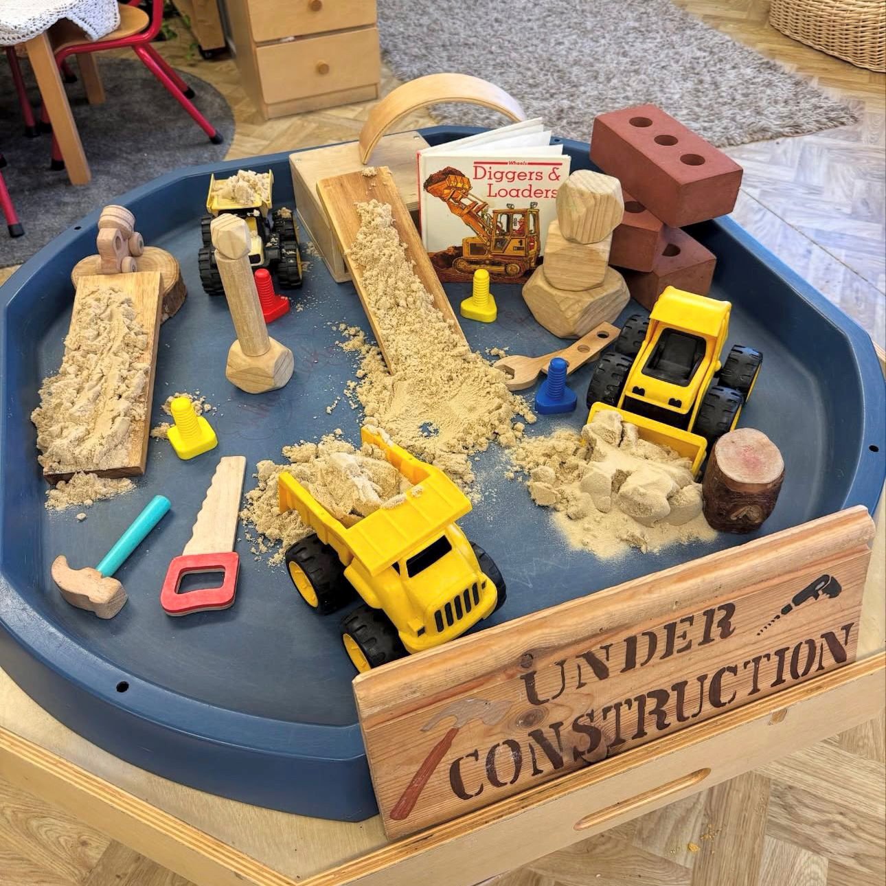 A children's sand and construction play table filled with toy construction vehicles, wooden blocks, sand, and tools.