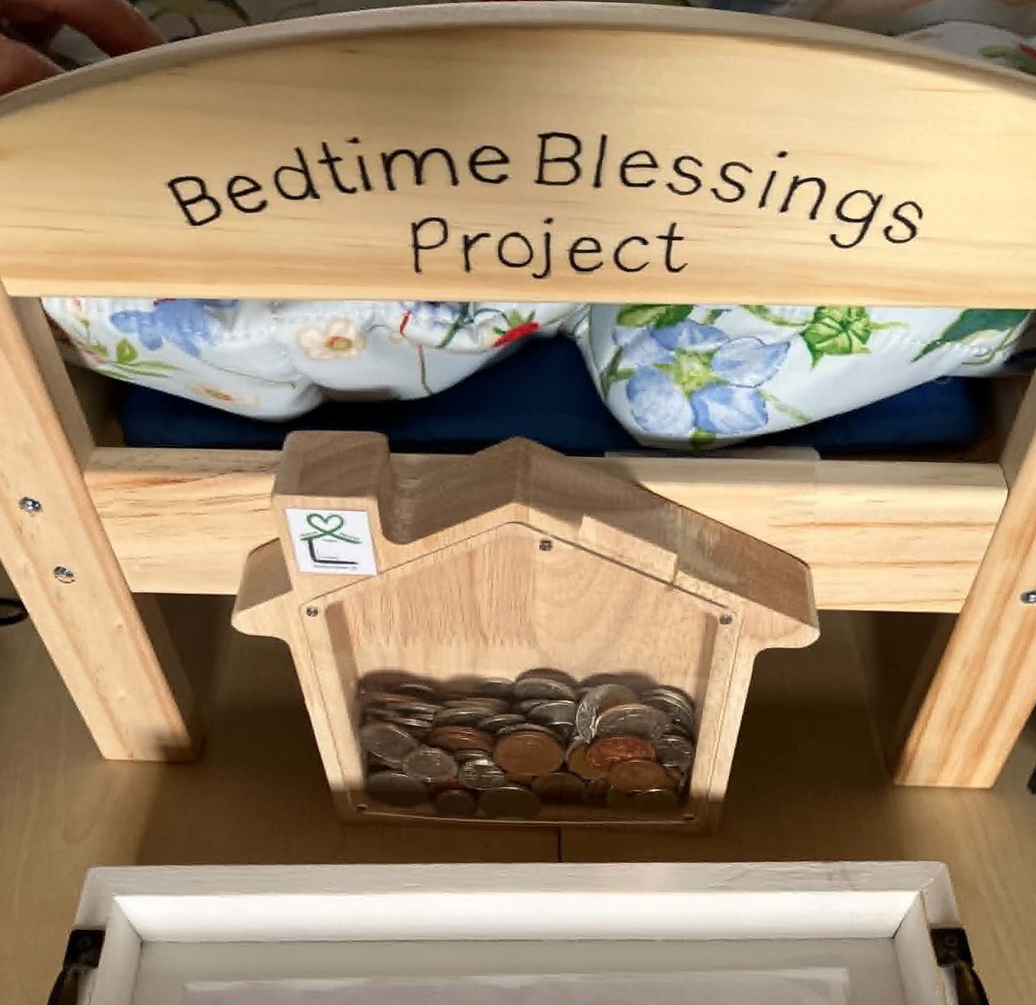 A wooden bed with a sign that reads 'Bedtime Blessings Project,' a blanket with floral patterns, and a small shelf with coins.