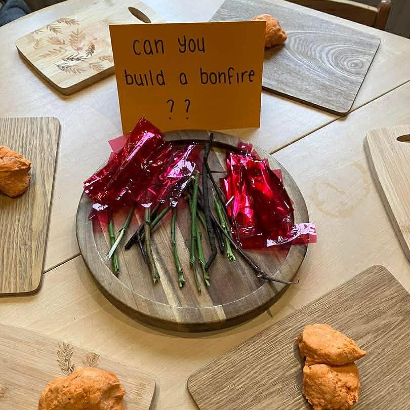 A round wooden board with sticks of red and green candy, resembling a fire with logs. A sign on the board says, "can you build a bonfire ??". Surrounding the table are various wood pieces and decorative trays.