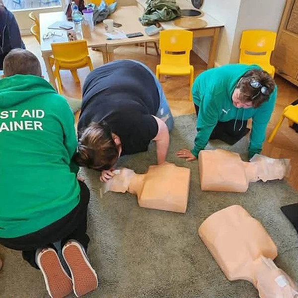 Three people practicing CPR on mannequins during a training session in a room with yellow chairs and a table.