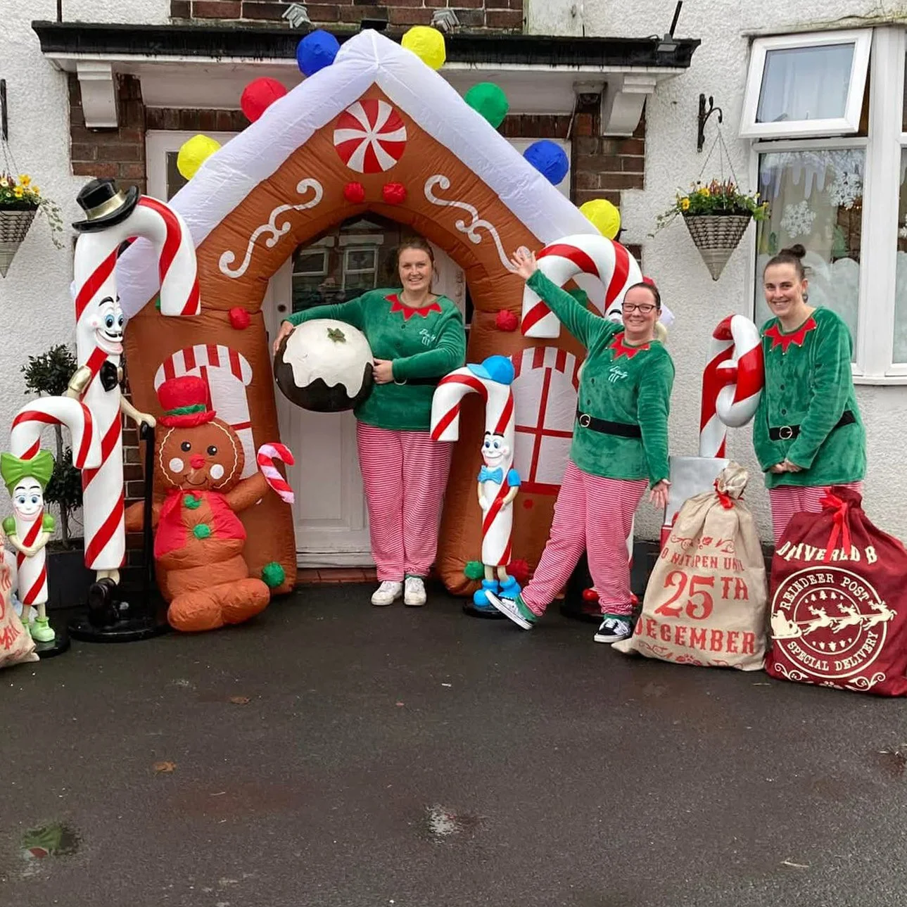 Three women dressed in Christmas-themed pajamas stand outdoors in front of a house with an inflatable gingerbread house and Christmas decorations. One woman holds a giant gingerbread cookie, and two are smiling and waving.
