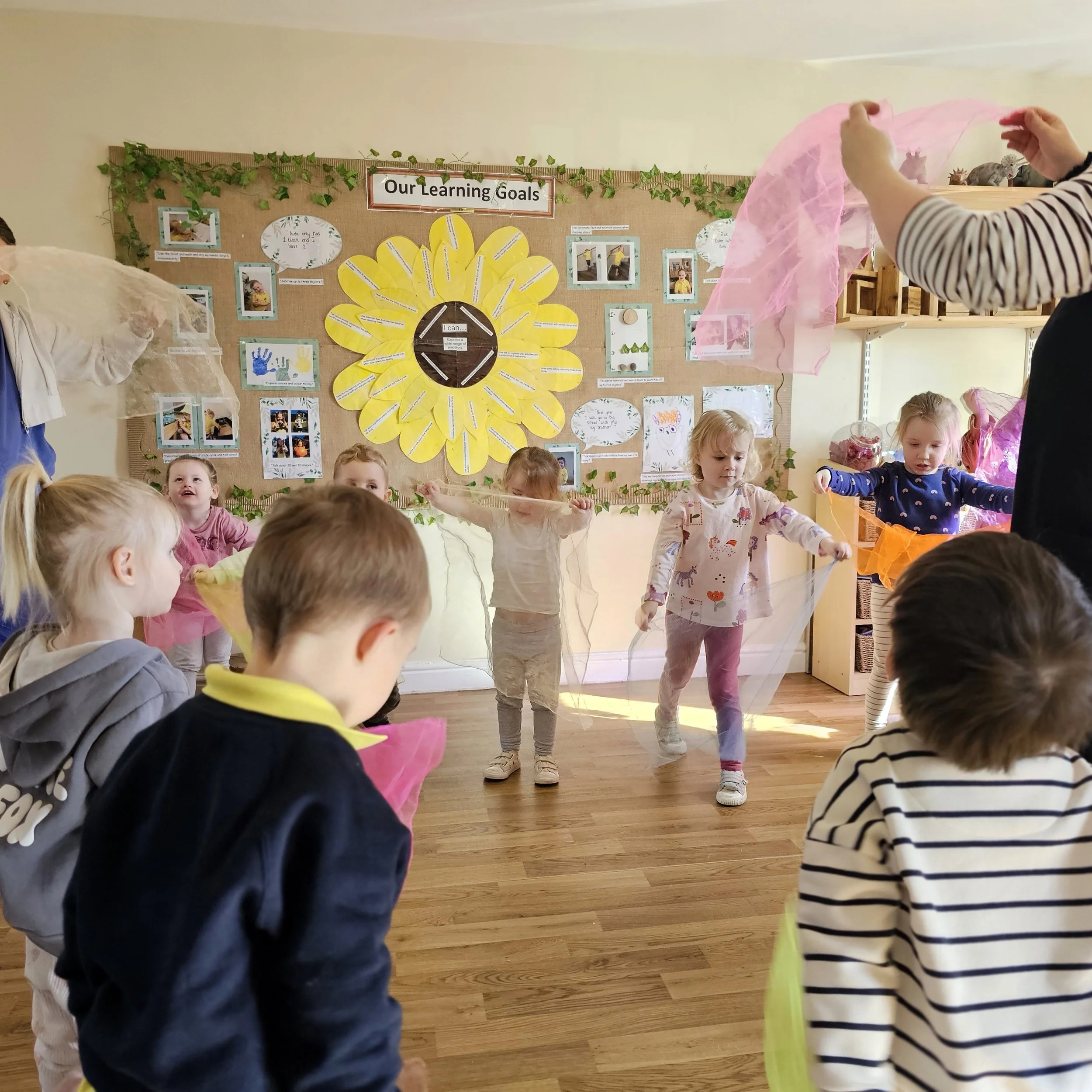 Children participating in a classroom activity involving colorful tissue paper and elastic bands, with a bulletin board titled "Our Learning Goals" in the background.