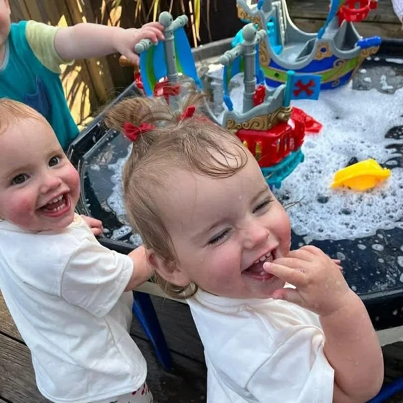 Two young children playing in a toy water table outdoors, with one child smiling and the other finger in her mouth, and colorful toy ships and accessories in the water.
