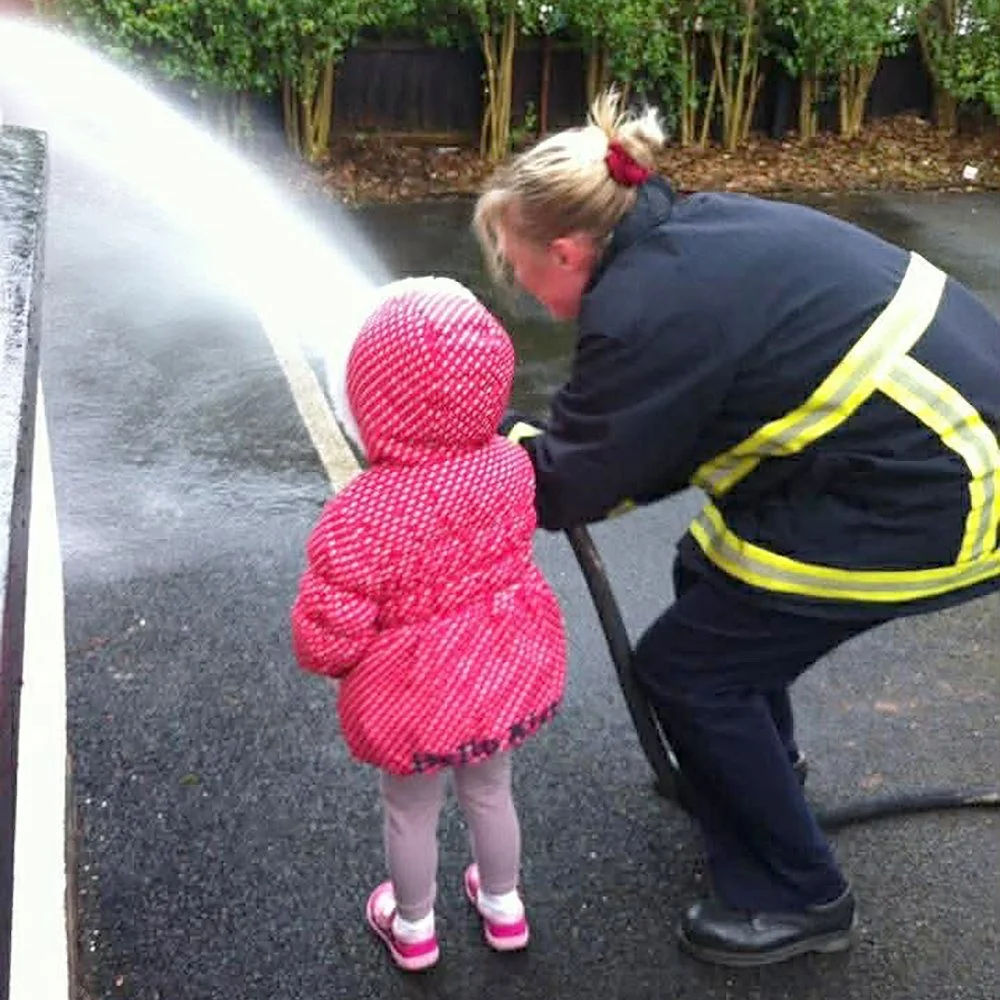 A firefighter helping a young girl learn how to use a fire hose on a wet street, with other greenery in the background.
