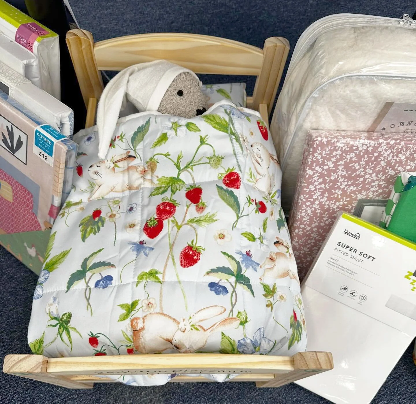 Child's bed with rabbit-themed bedding featuring strawberries, flowers, and rabbits, set among household items like mattress protectors, books, and a green box.
