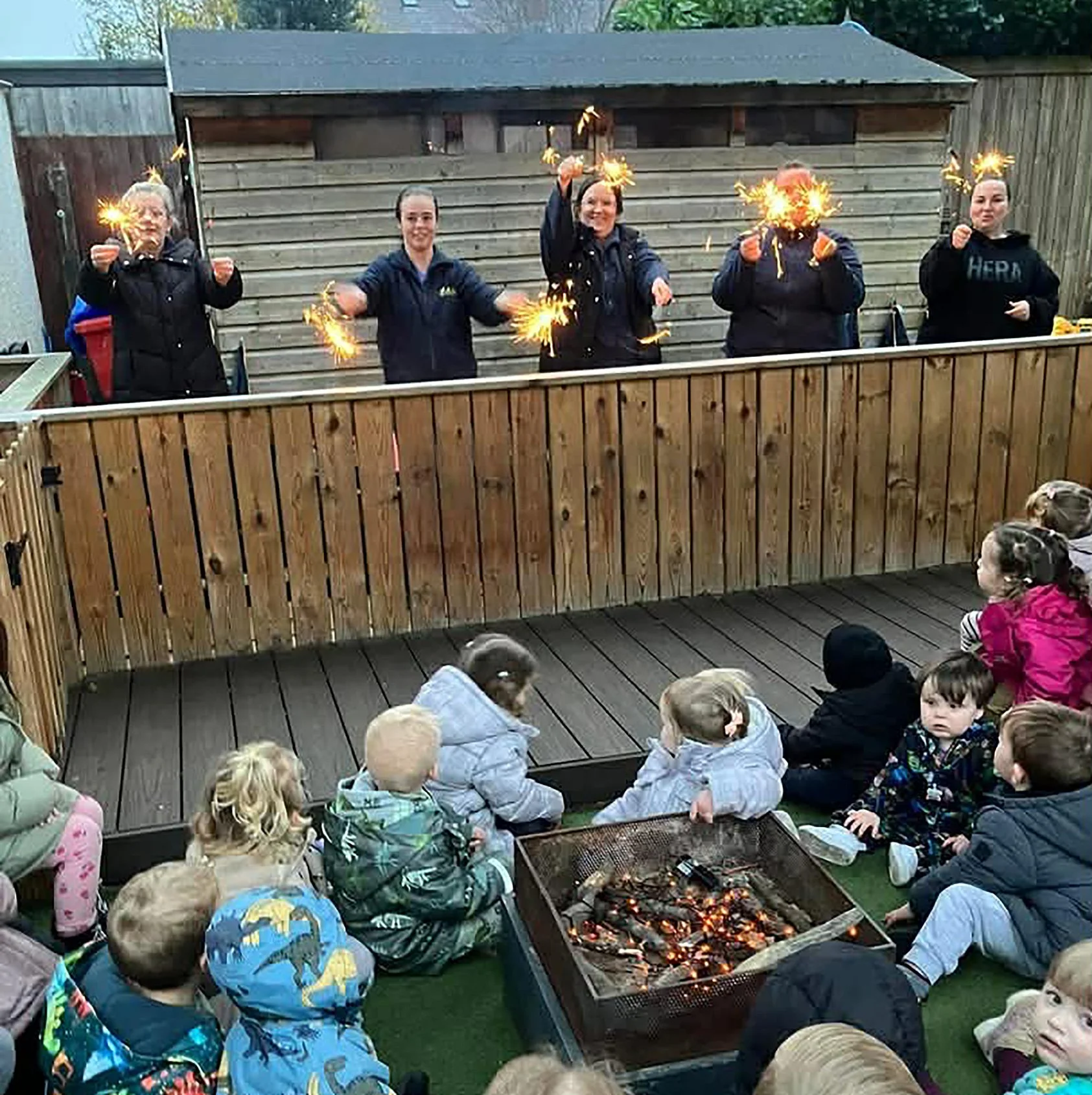 Children and adults sitting around a fire pit outdoors, watching a celebration where five women are holding sparklers in front of a wooden shed, with children seated on the ground in front.