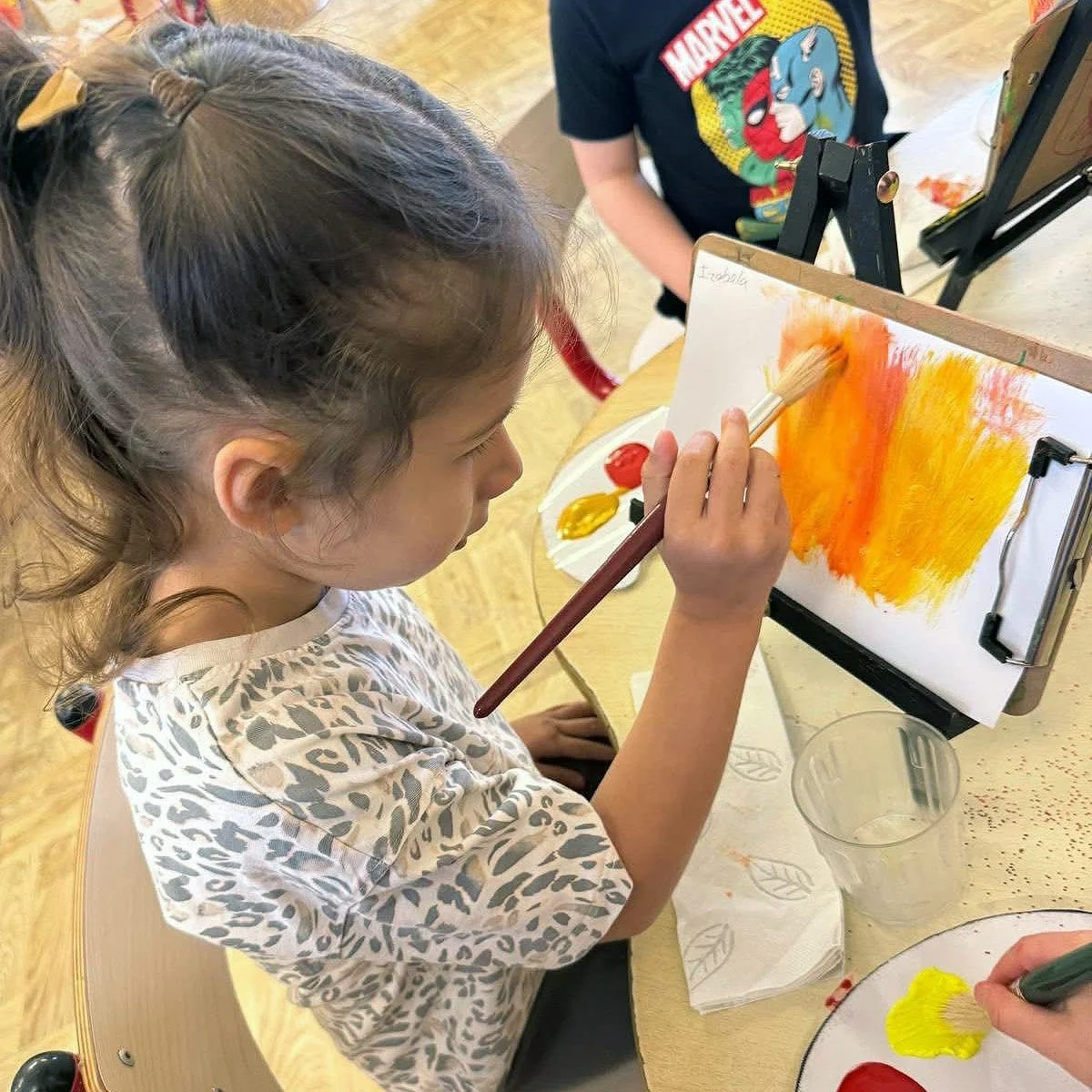 A young girl painting with a brush on a small easel, creating an abstract orange and yellow background. She is seated at a table with a palette of red and yellow paint, and nearby are a glass of water and a plate with yellow paint.