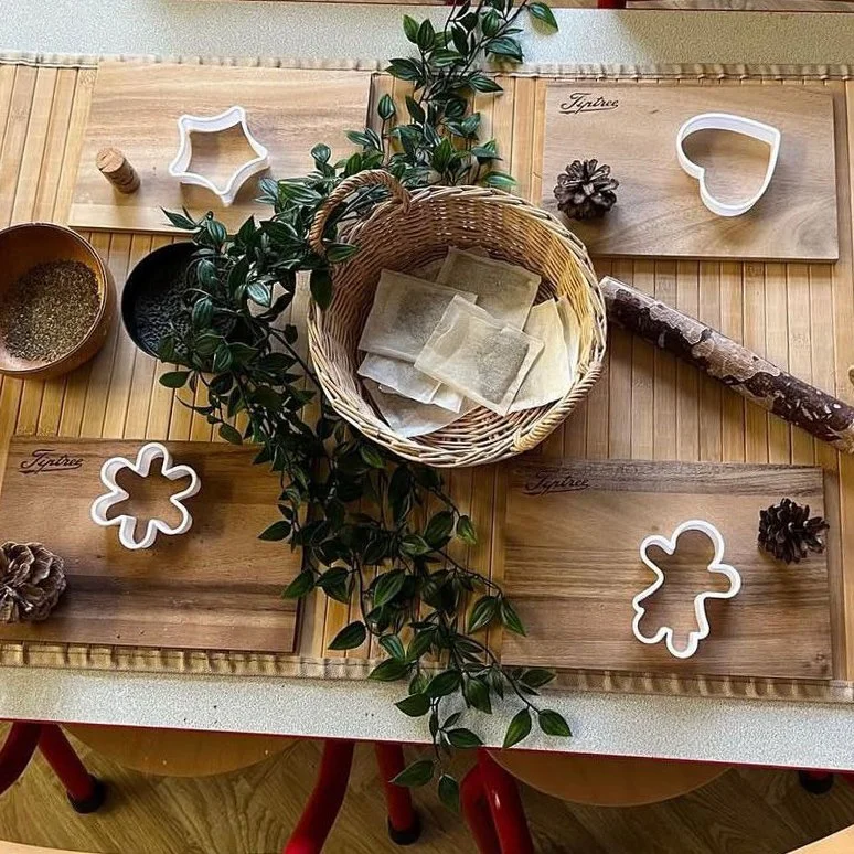 A crafting table with wooden boards, cookie cutters, pinecones, a basket with parchment paper, a sprig of greenery, and a rolling pin, set up for holiday baking and decorating.