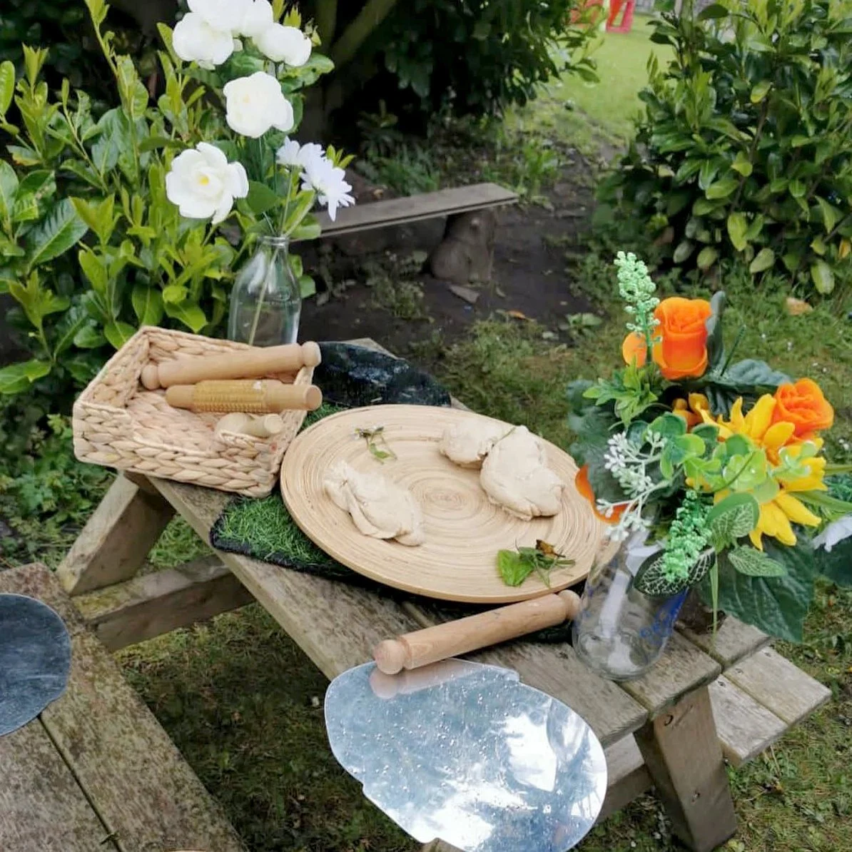 An outdoor scene showing a rustic wooden table with a flower arrangement, a basket with wooden rolling pins, a wooden cutting board with dough, a rolling pin, and metal pie tins. There is a glass vase with white flowers and greenery, and a small bench in the background among green bushes.
