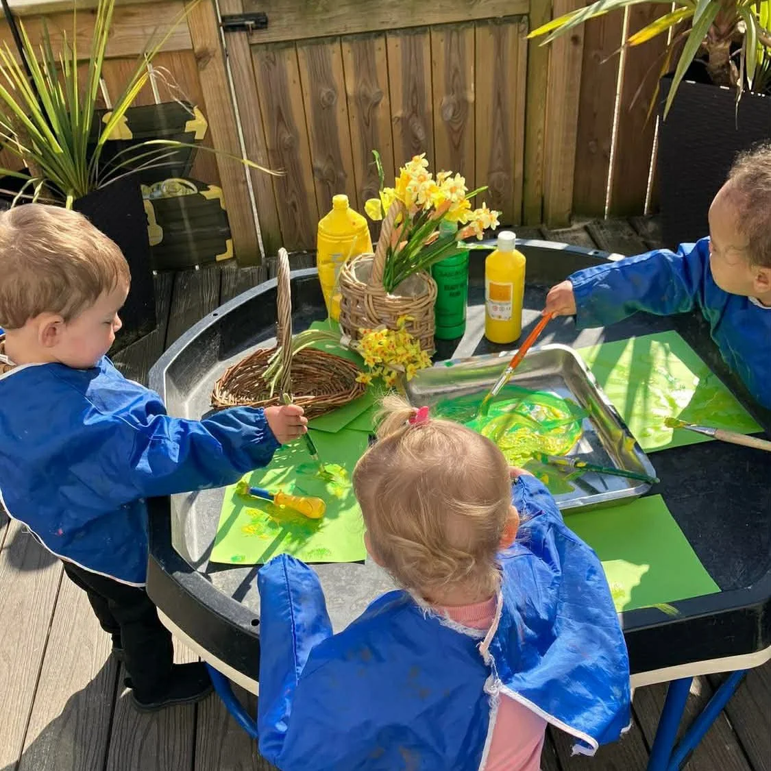 Three young children painting with yellow and green colors at an outdoor table, surrounded by potted plants, baskets of daffodils, and bottles of yellow paint.
