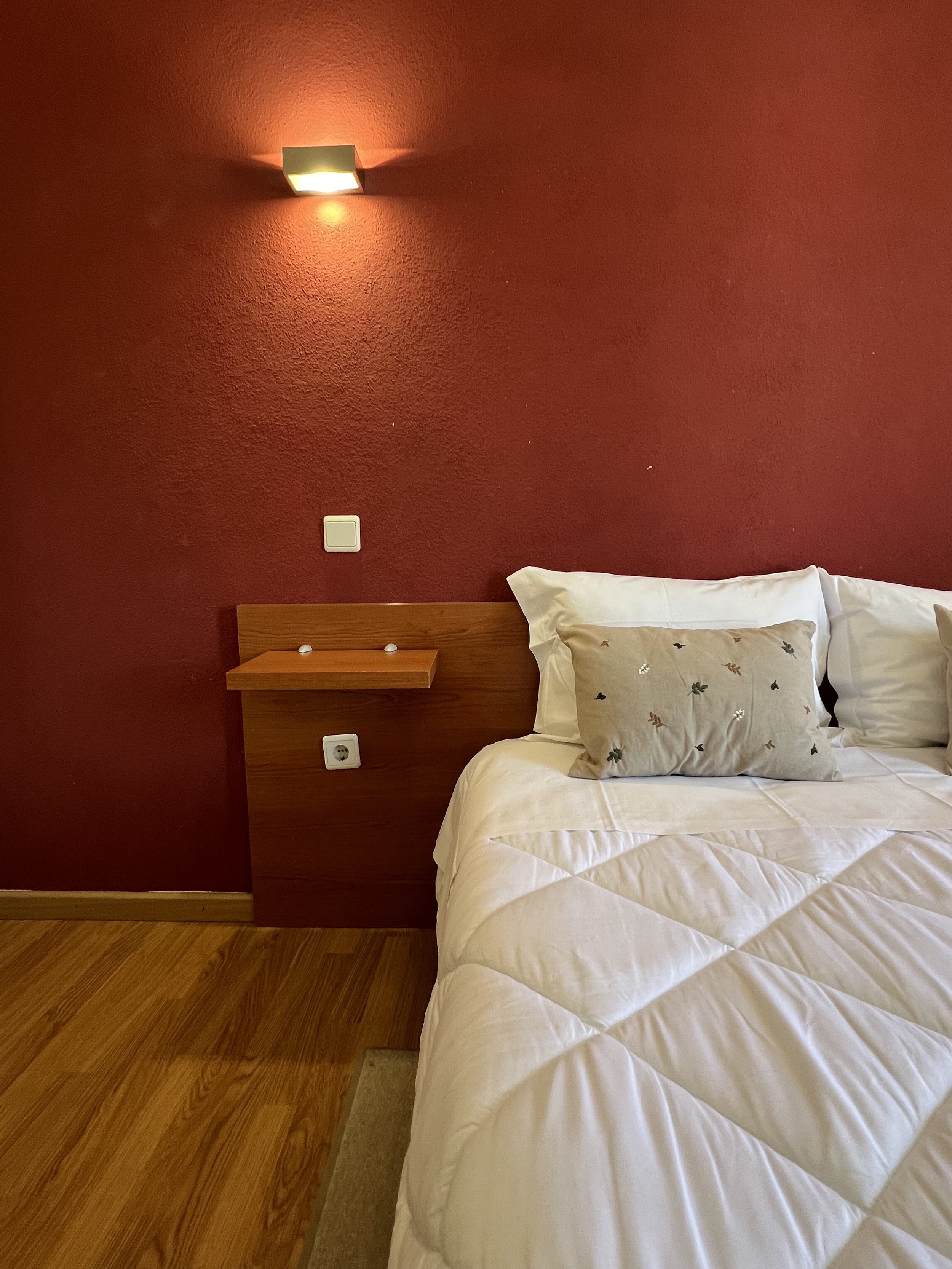 A hotel room with a bed, white bedding, and pillows, along with a red wall, wooden headboard, side table, wall-mounted light fixture, and electrical outlets.