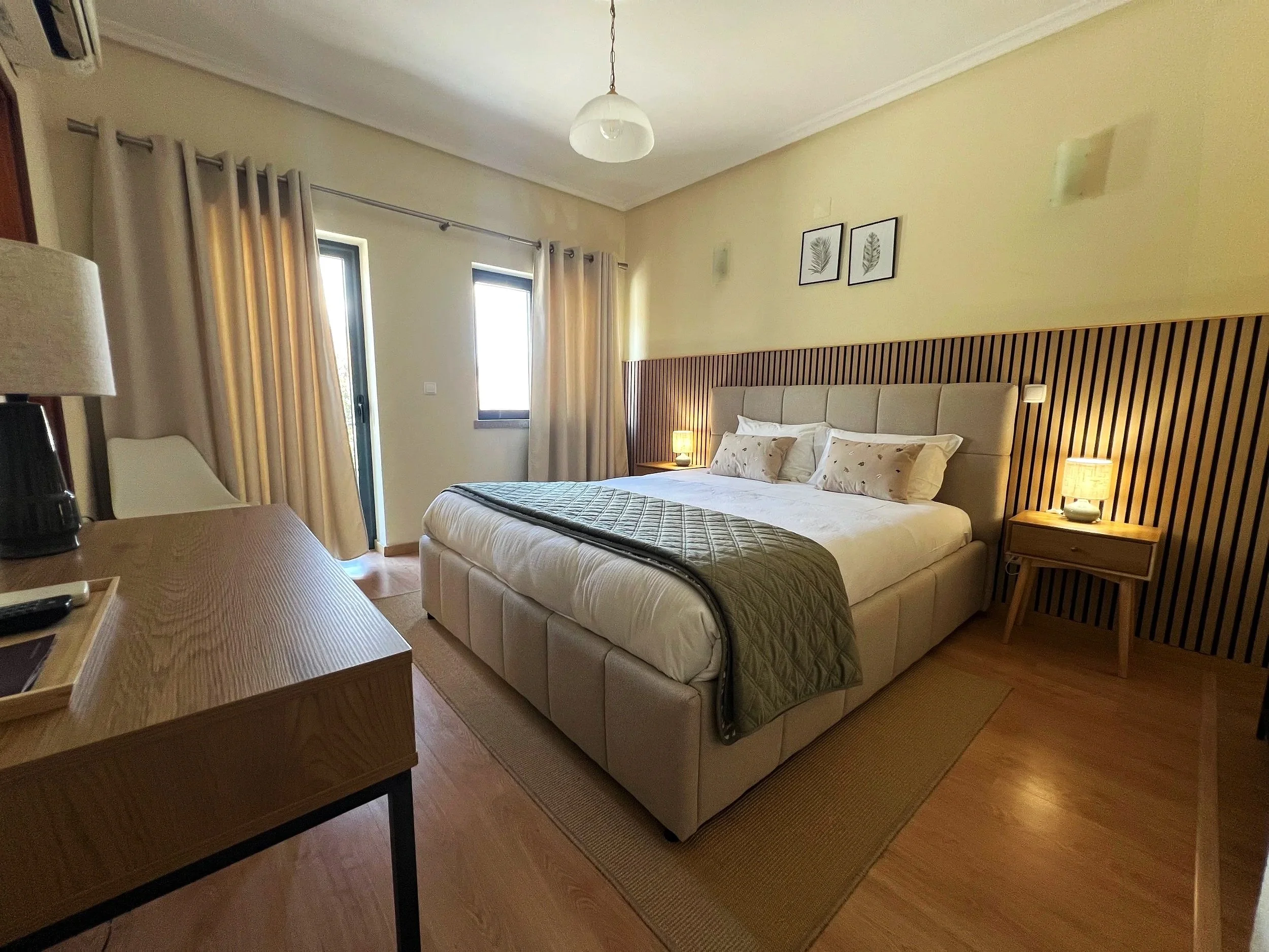 A bedroom with a large bed, framed prints of leaves above the headboard, bedside lamps, windows with curtains, and a wooden desk with a lamp and remote controls.