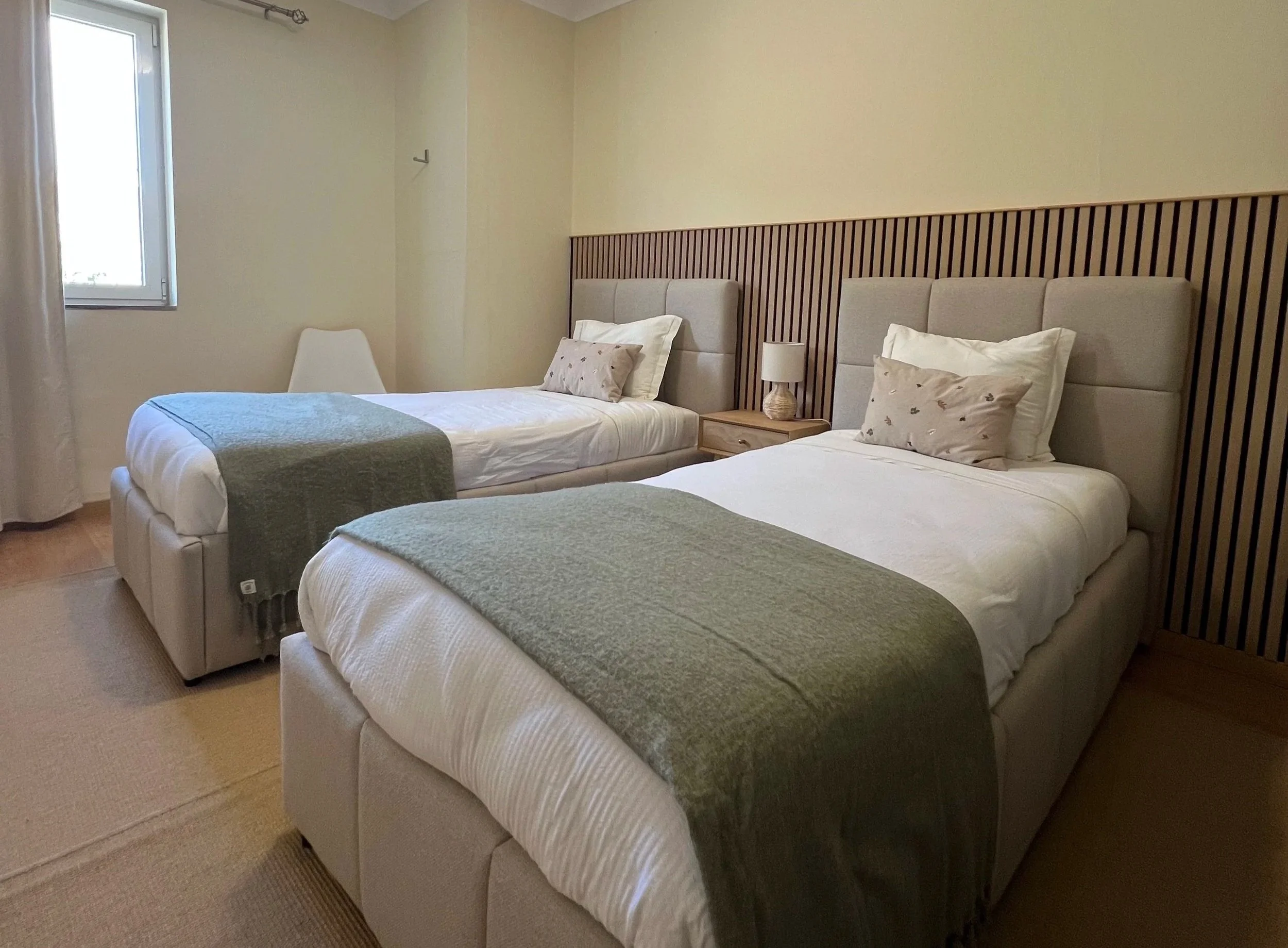 A hotel room with two beds featuring beige upholstered headboards, pillows, and green blankets. There is a nightstand between the beds with a small lamp, and a chair near the window with beige curtains and a window with white framing.