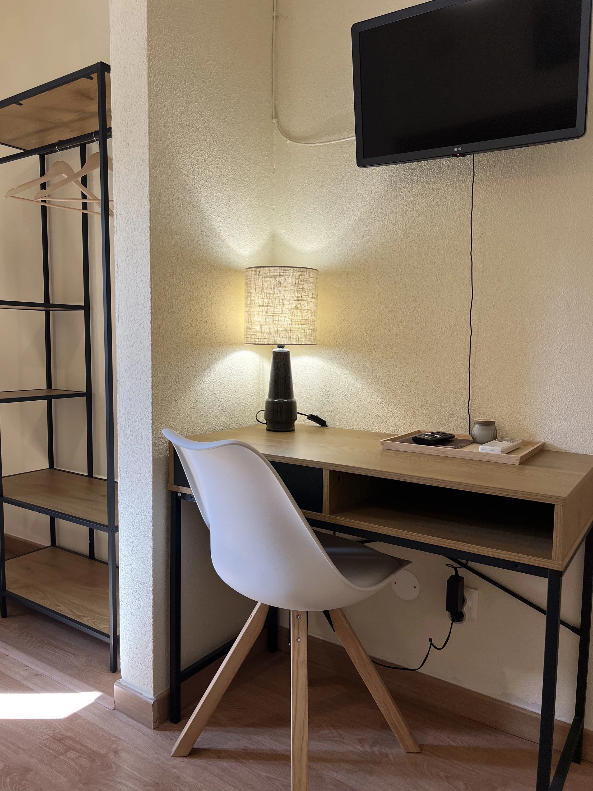 A workspace with a wooden desk, a modern white chair, a tall table lamp, a flat-screen TV mounted on the wall, and a black metal and wood shelving unit.