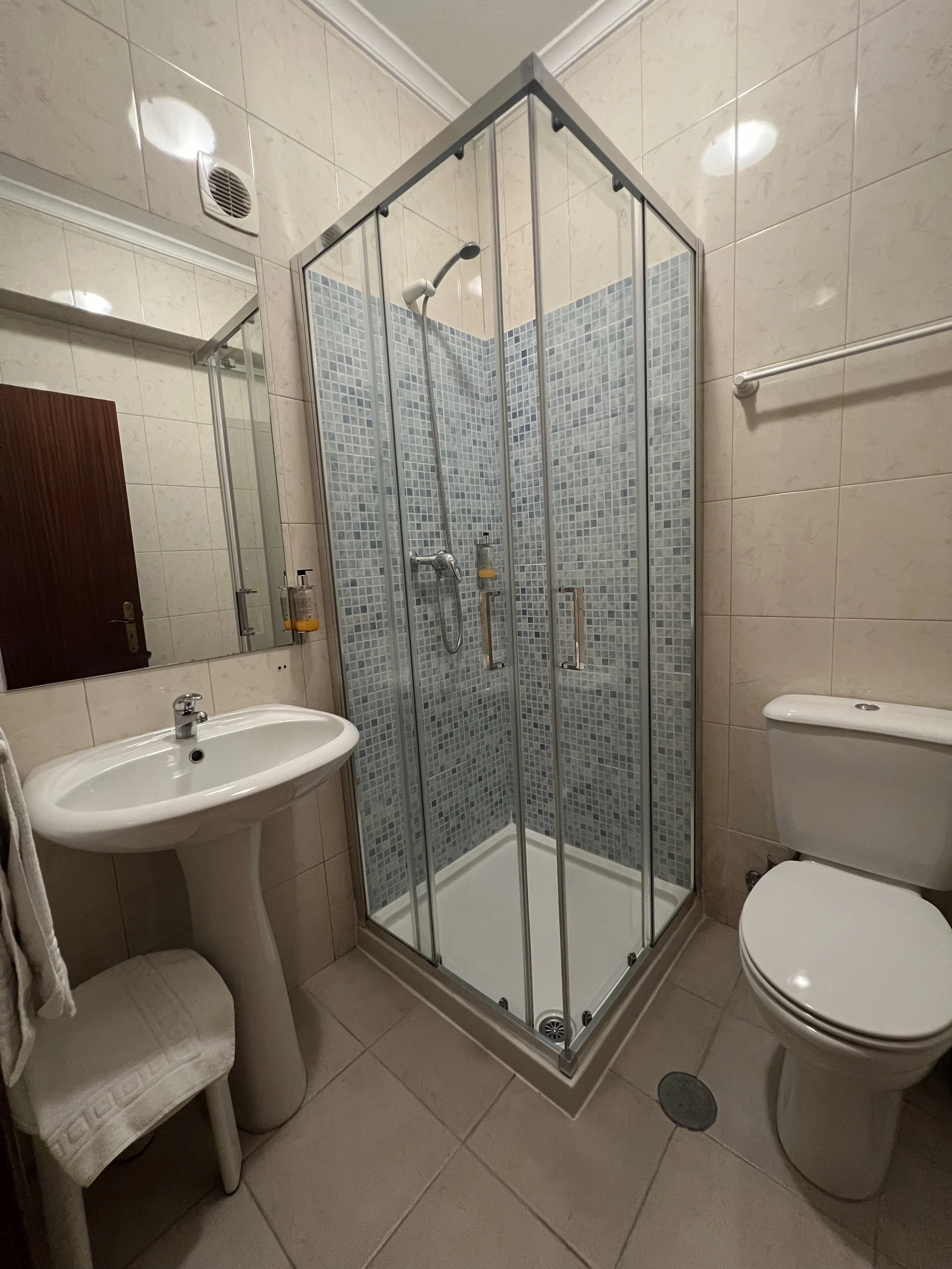 Bathroom with a shower enclosure, a white toilet, a small white sink, mirror, and beige tiled walls and floor.