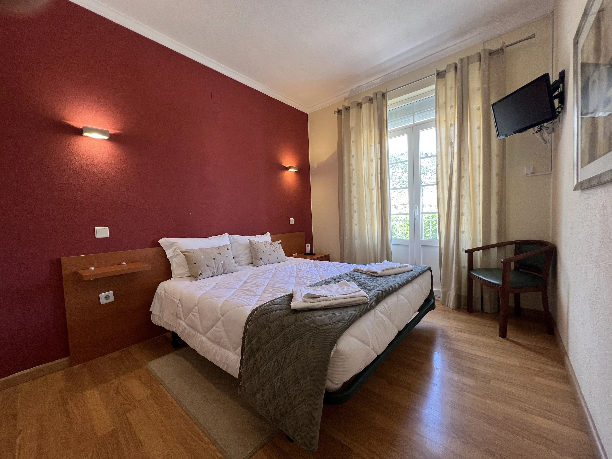 A bedroom with a double bed, white bedding, and two pillows. The headboard is made of wood with attached small shelves. The wall behind the bed is painted dark red, with two wall-mounted lights. There is a window with light-colored curtains, a small black flat-screen TV mounted on the wall, and a wooden chair with a dark green cushion beside the window. The floor is wooden.