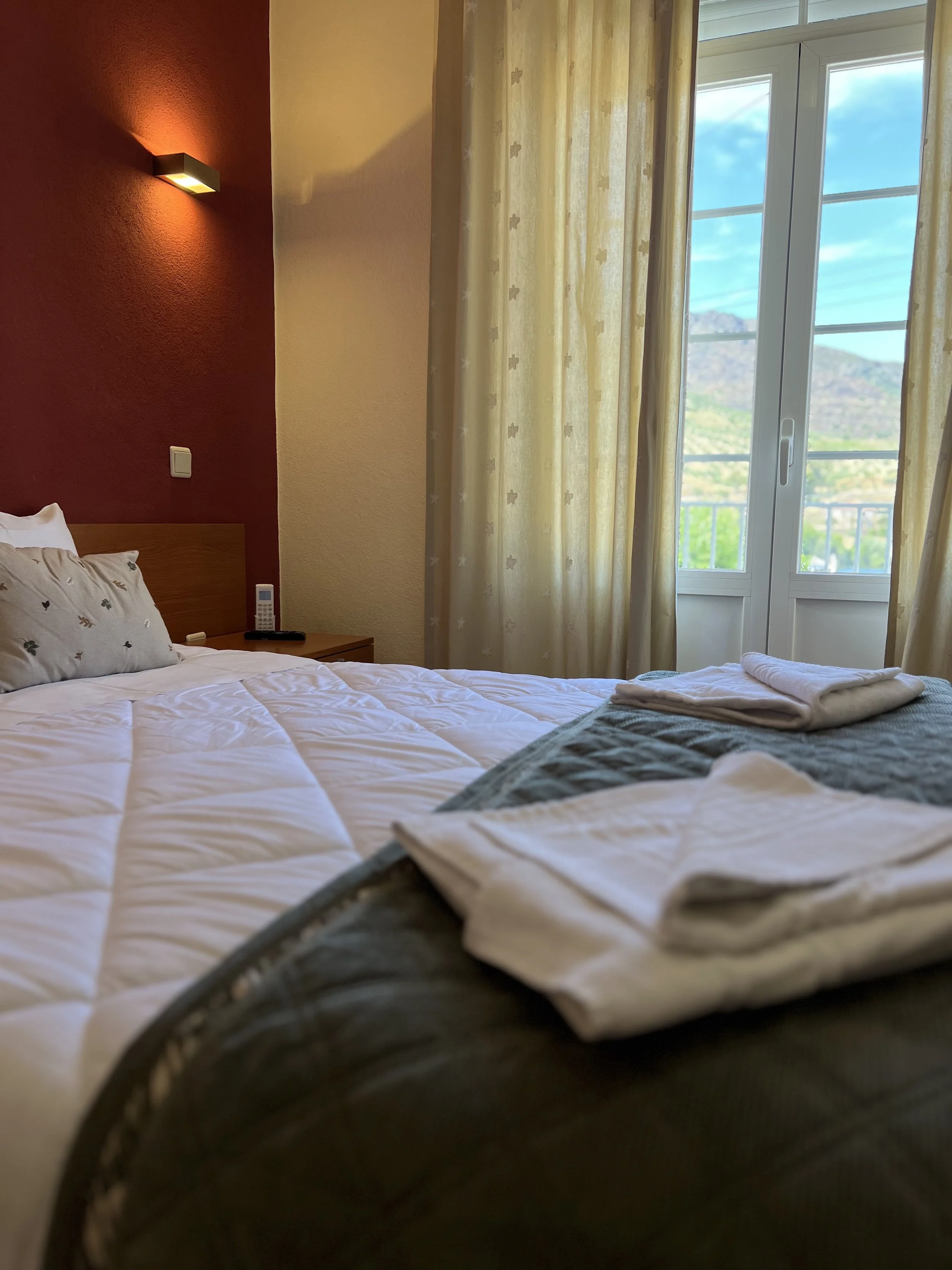 A neatly made bed with white sheets, a gray bed runner, and folded white towels on top, in a hotel room with a large window and yellow curtains, outside mountain scenery visible.