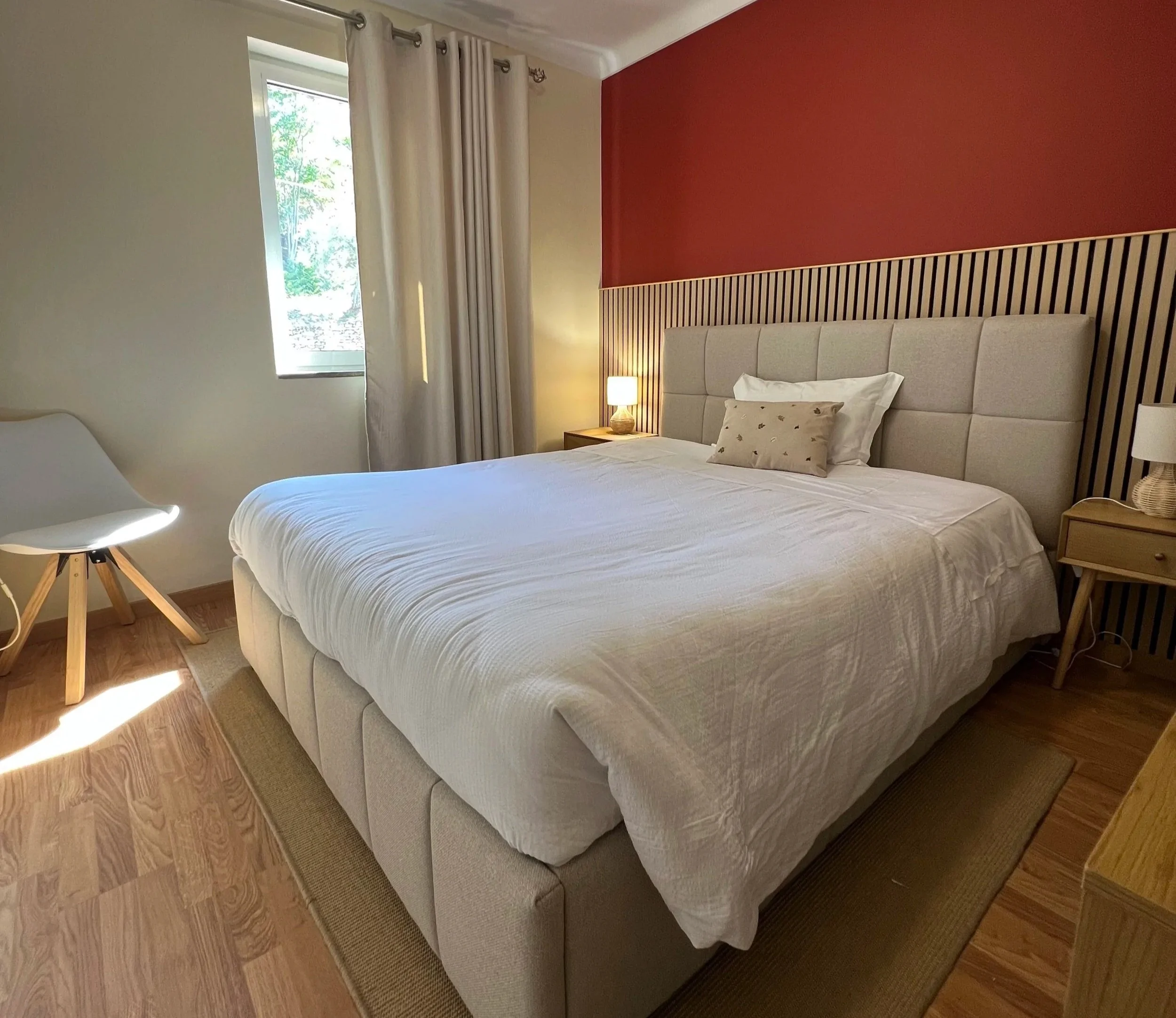 A bedroom with a large bed featuring a light-colored upholstered headboard, white bedding, and pillows. A small nightstand with a lamp is beside the bed. There is a window with beige curtains and wood flooring.