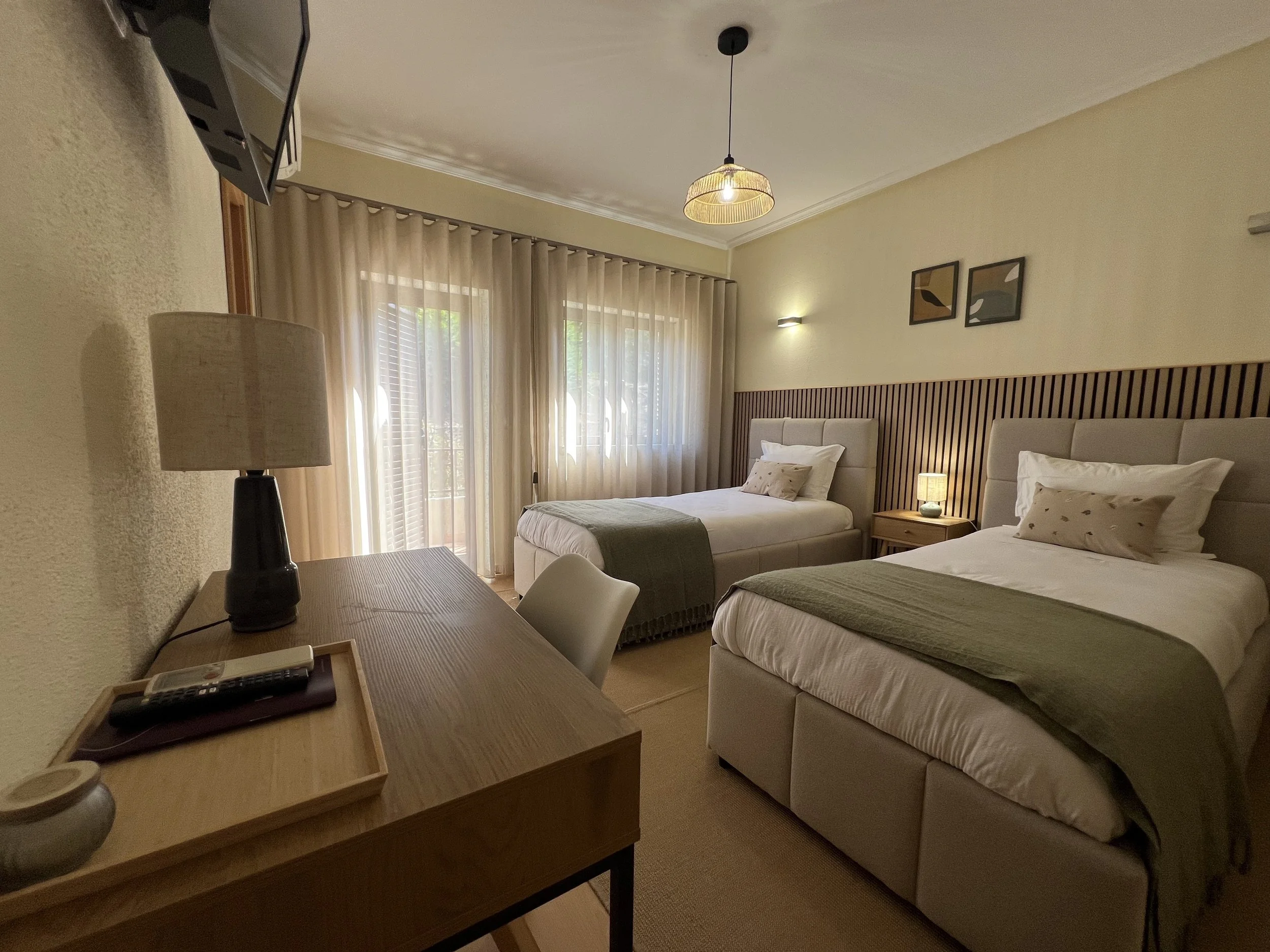 A hotel room with two beds, beige walls, curtains, a table with a lamp, and a television mounted on the wall.