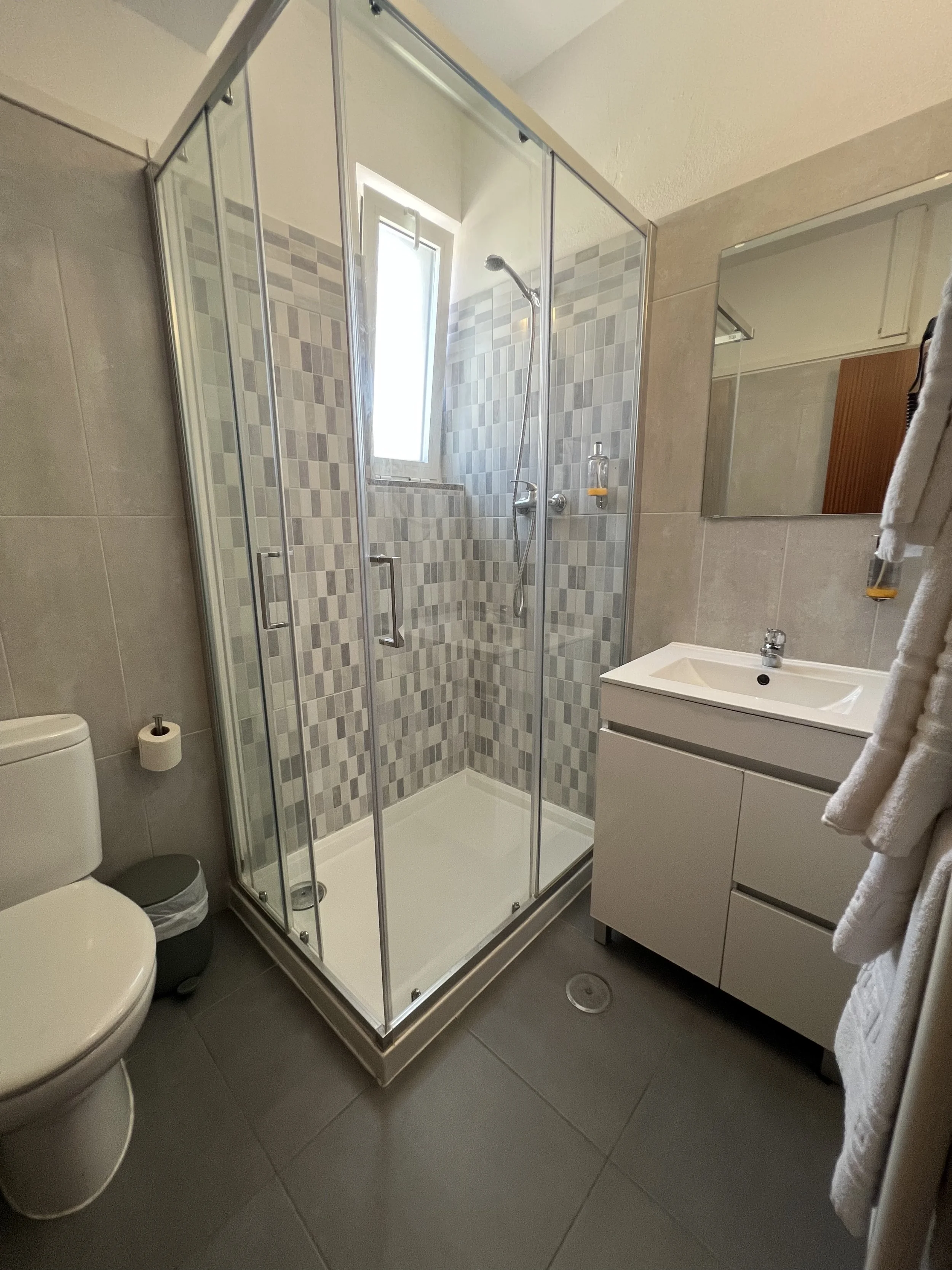 A modern bathroom with a walk-in shower featuring glass doors and tiled walls, a white vanity with a sink, a mirror, a toilet, and bathroom accessories.
