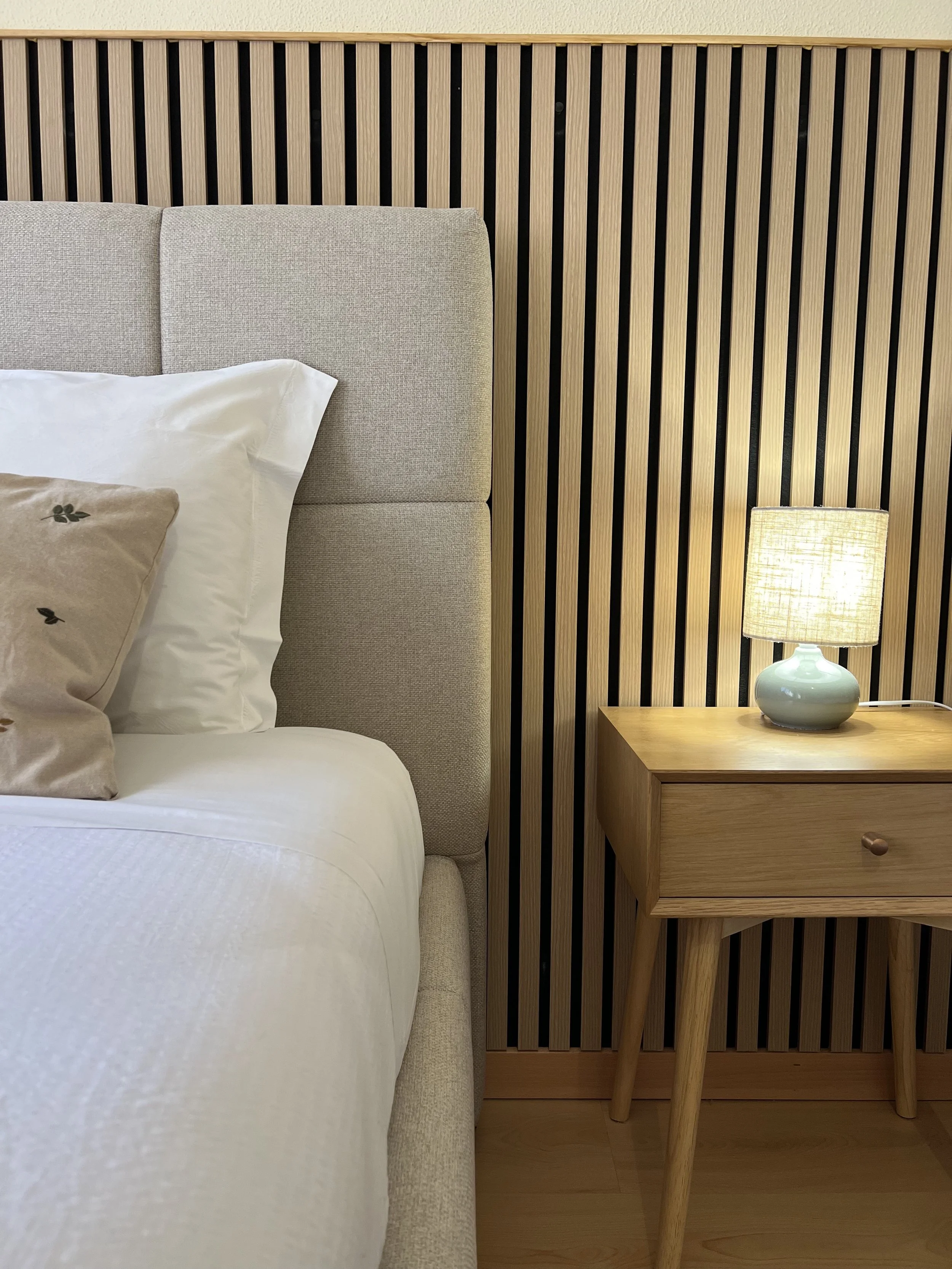 Side view of a bed with beige upholstered headboard, white pillows, and a light wood nightstand with a ceramic table lamp.