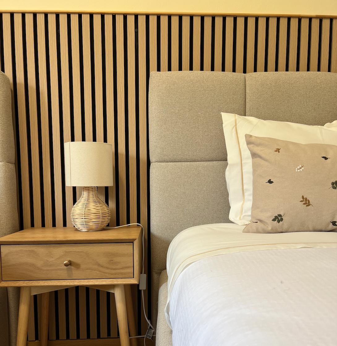A bedroom with a beige upholstered headboard, a wooden nightstand, a wicker table lamp, and a pillow with a botanical print on a white bedspread. The headboard extends to the right side of the image, and the background features a vertical striped wooden panel.