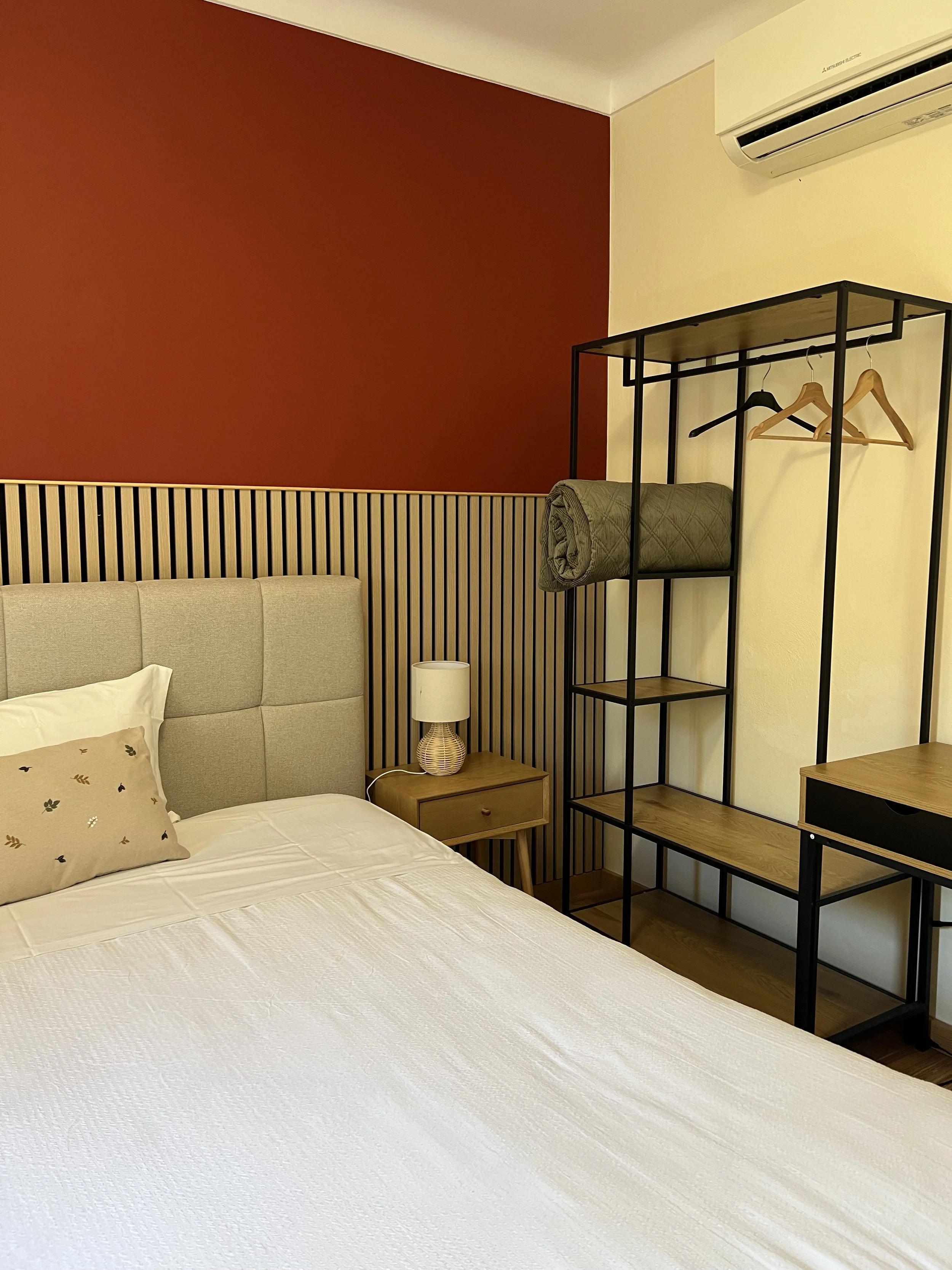A bedroom with a bed, beige headboard, white bedspread, decorative pillow, small nightstand with lamp, and a black metal open wardrobe with hanging space, rolled blanket, and wooden shelves. The wall behind the bed has beige and rust-colored sections, with a wooden slat accent.