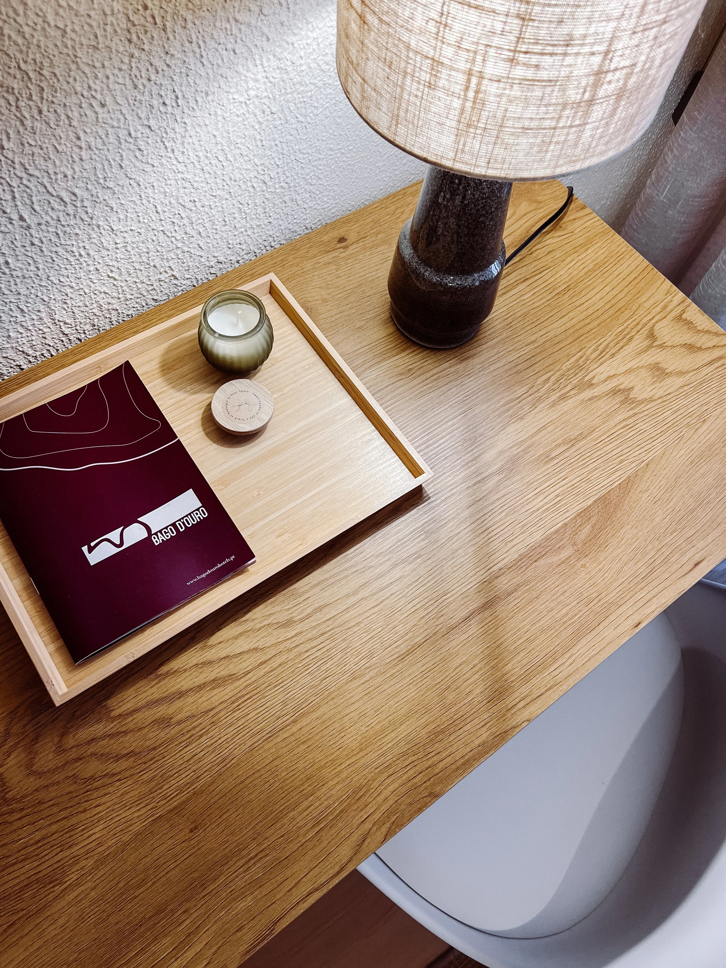 A wooden surface with a tray containing a candle in a glass holder, a small round container, and a burgundy brochure or magazine with the words "Bago Douro" and a logo on it. There is a table lamp with a beige lampshade on the right side of the surface, and part of a washing machine or dryer is visible underneath the table.