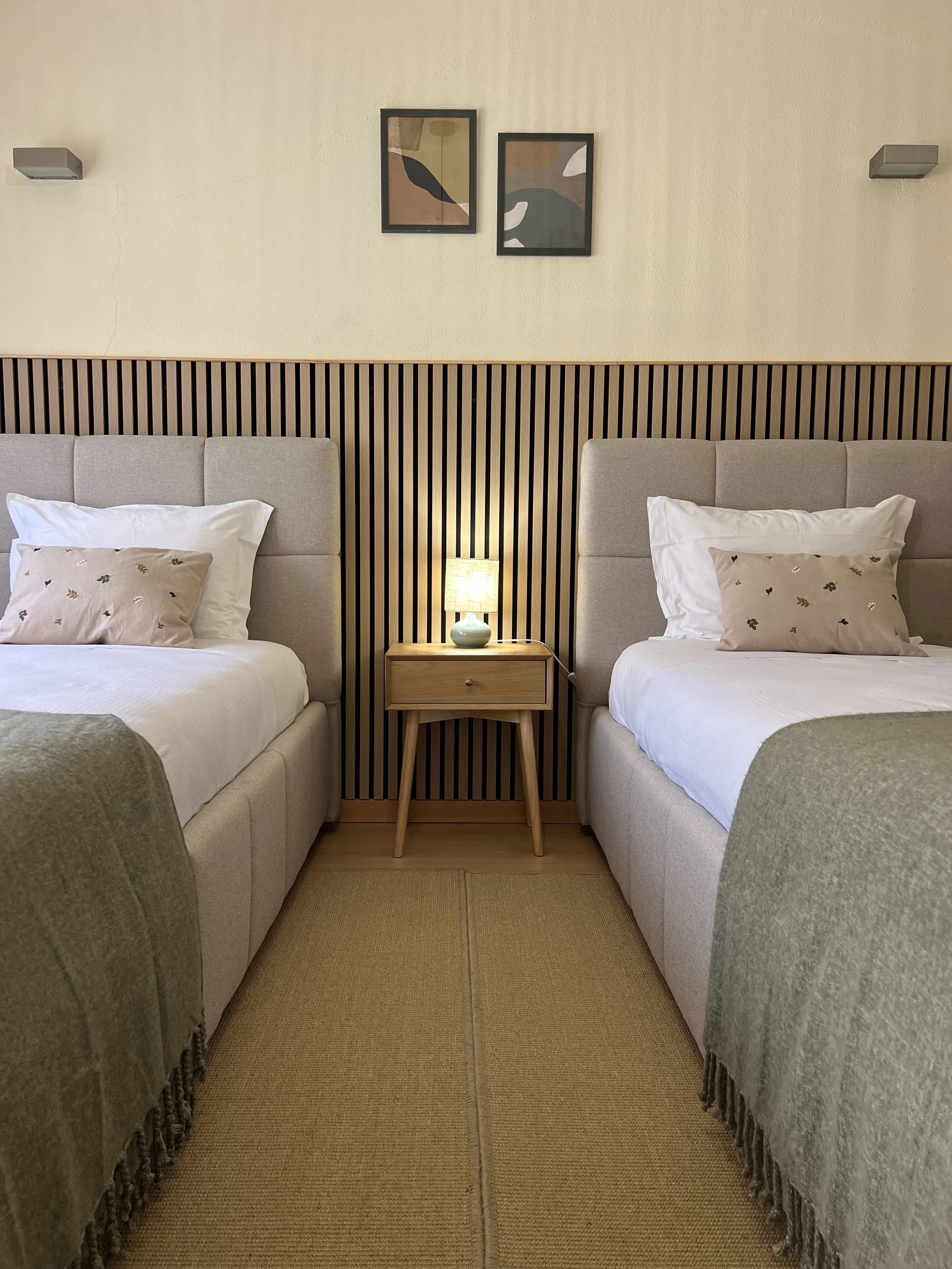 A bedroom with two twin beds separated by a nightstand with a small lamp. The beds have beige headboards, patterned pillows, and white bedding. The wall behind the beds has a wooden slat panel and two framed abstract artworks. The floor has a woven beige rug.