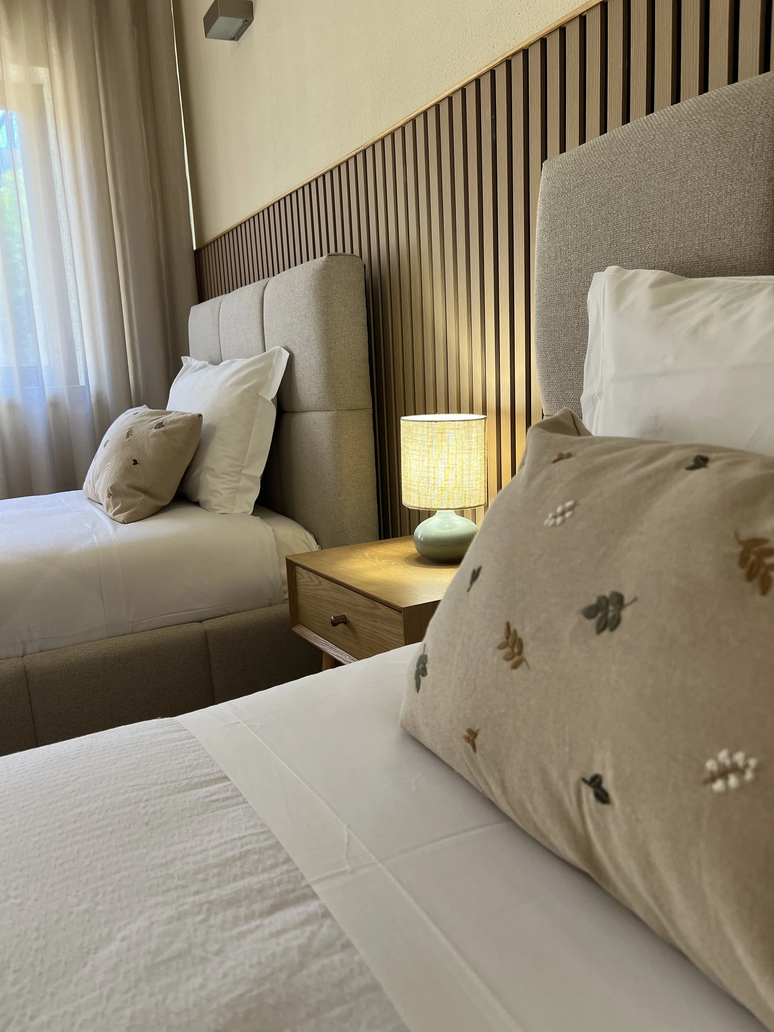 Two beds with beige headboards and white bedding in a hotel room, with a wooden nightstand, a yellow lampshade, and curtains over a window.