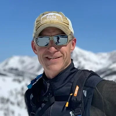 Man taking a selfie in snowy mountain landscape, wearing sunglasses, a baseball cap, and outdoor gear.