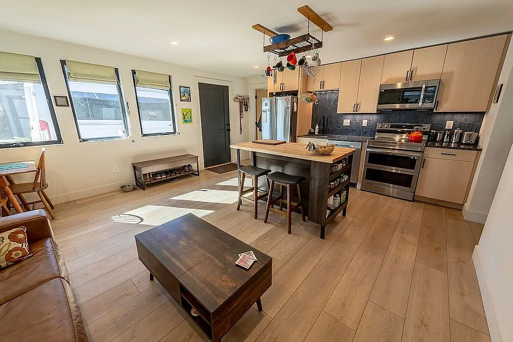 Open-concept kitchen and living area with hardwood floors, light wood cabinetry, stainless steel appliances, a kitchen island with stools, and windows with green blinds.