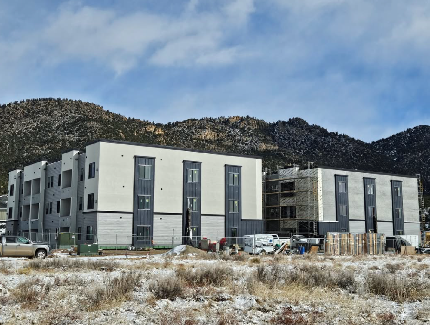 Construction site with modern apartment buildings, some finished and others under construction, with a mountain range in the background, parked vehicles, and construction materials in the foreground.